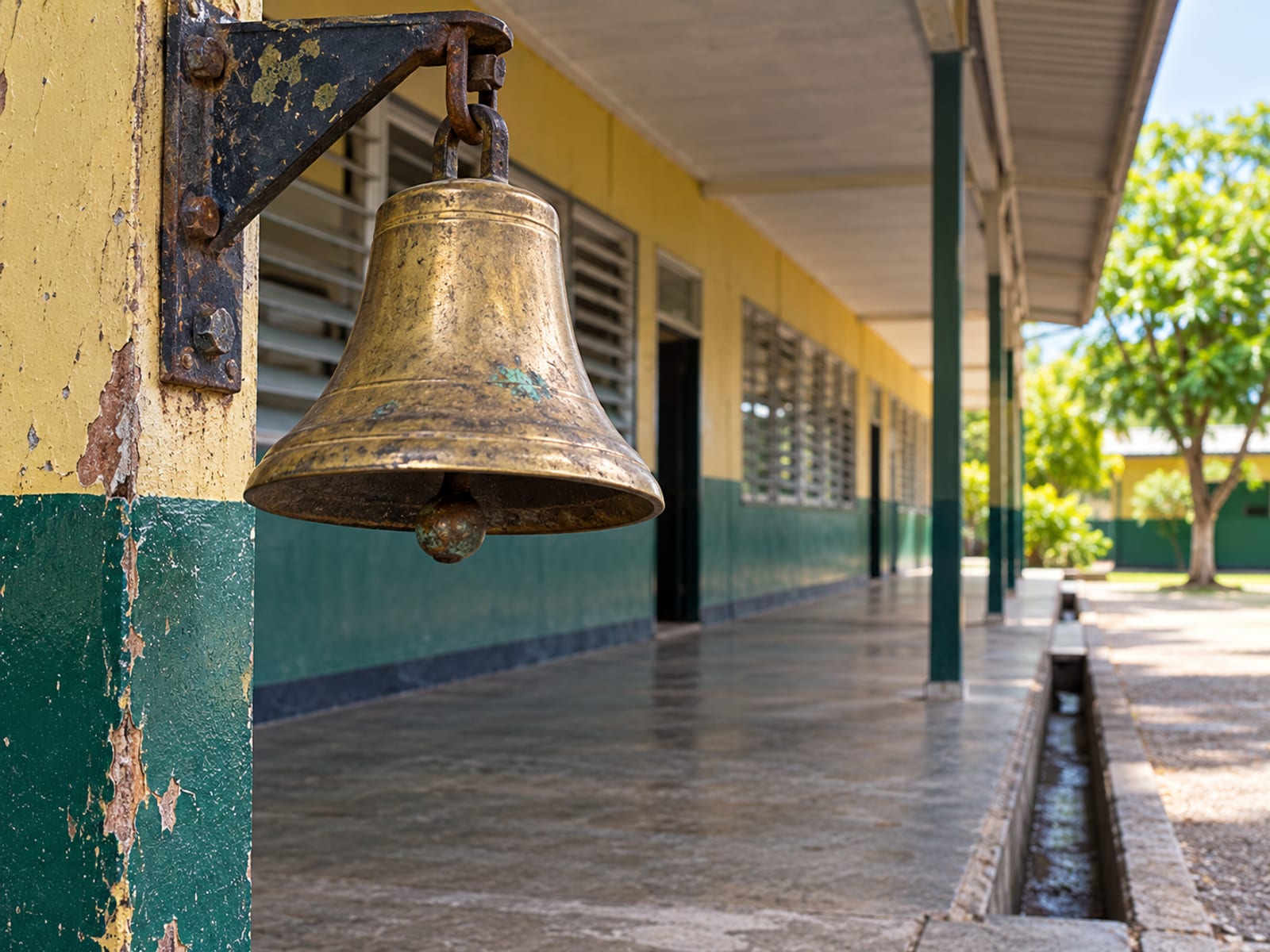 Brass school bell hanging from a post on a covered walkway, louvre classroom windows blurred behind