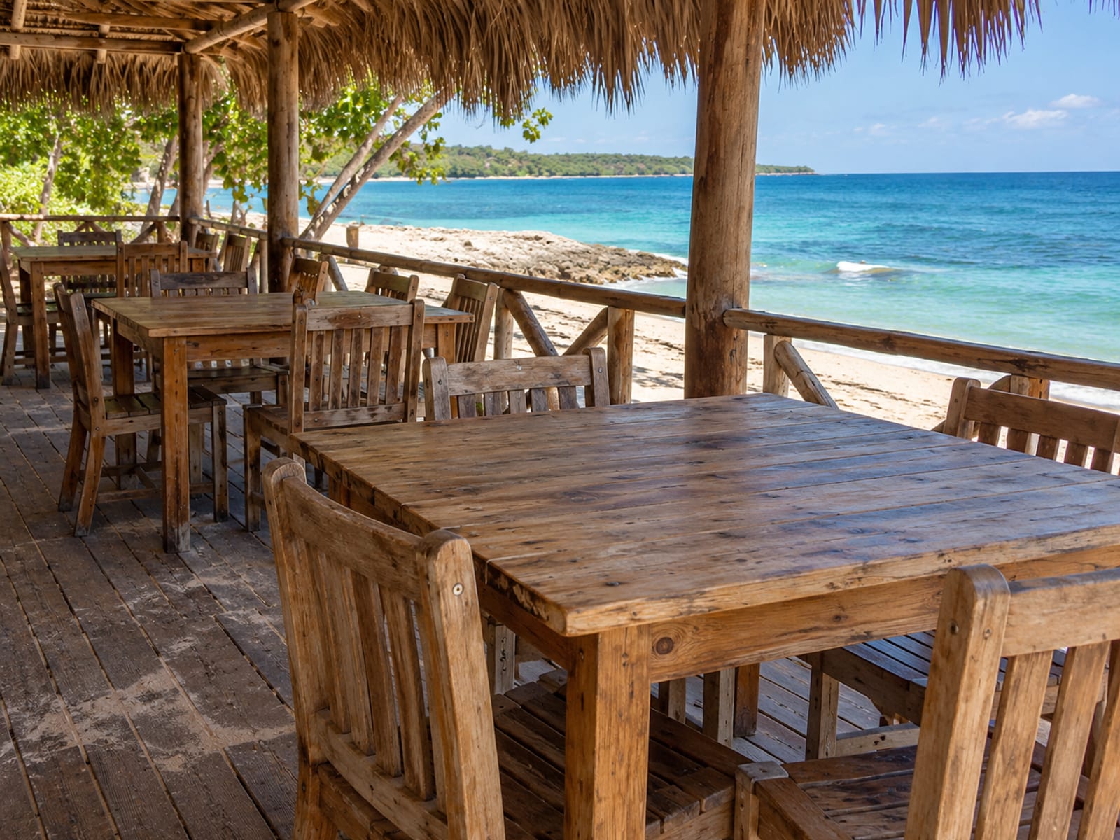 Rows of plain wooden tables and chairs on an empty open terrace beside the sand and sea.