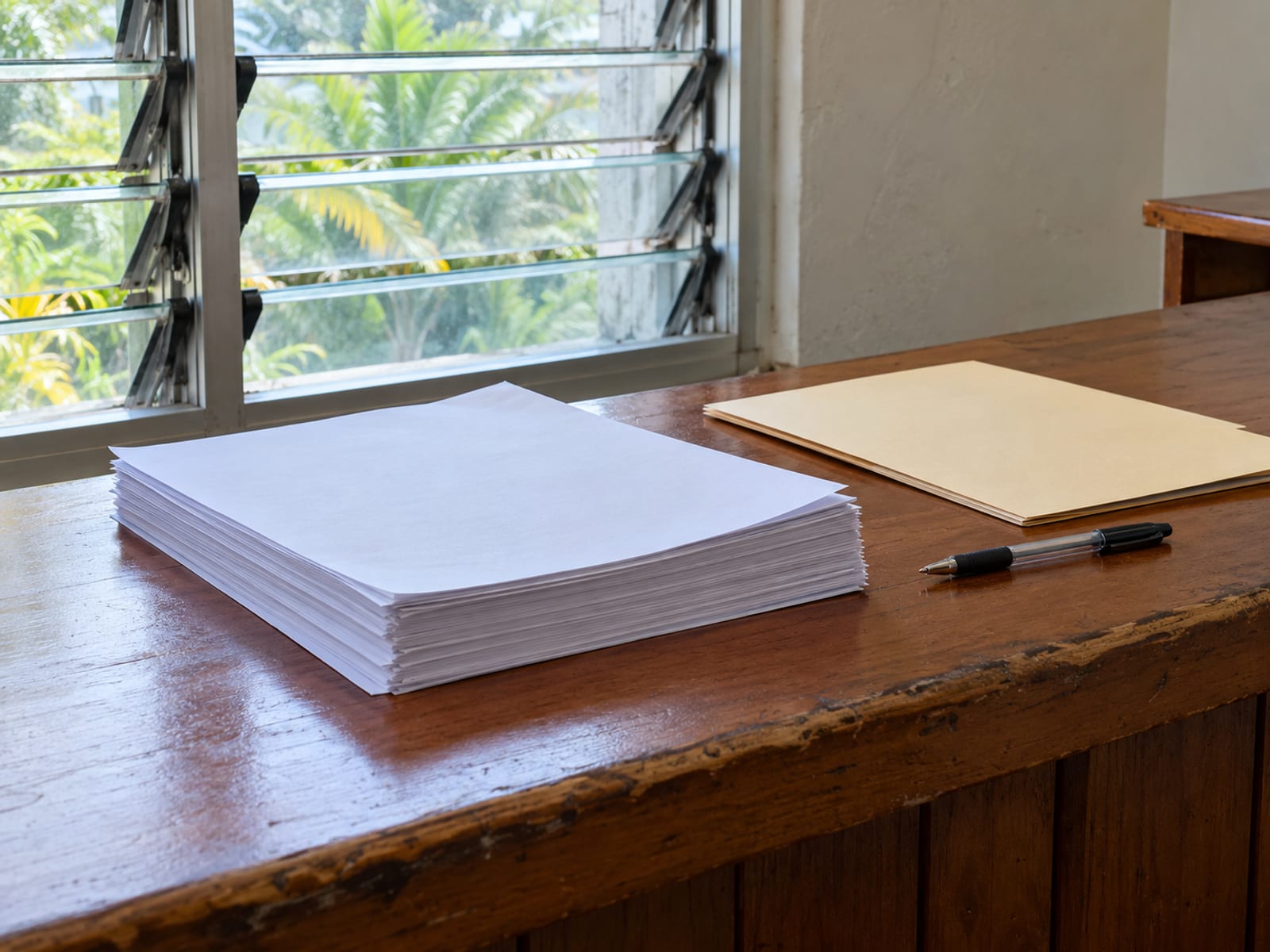 Wooden office counter with stacked blank sheets of paper, a closed folder and a pen, with a louvre window behind.