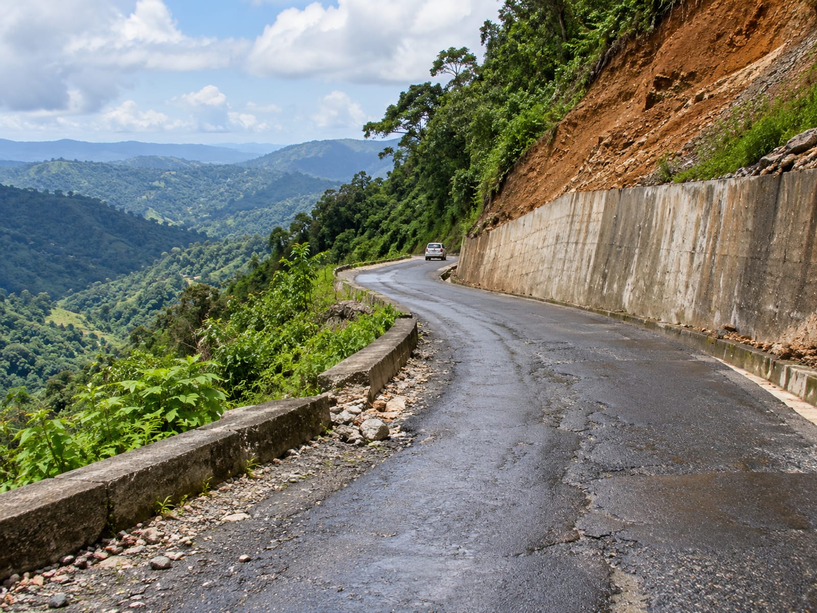 Narrow paved mountain road curving along a steep bank with a concrete retaining wall and a patch of slipped earth.