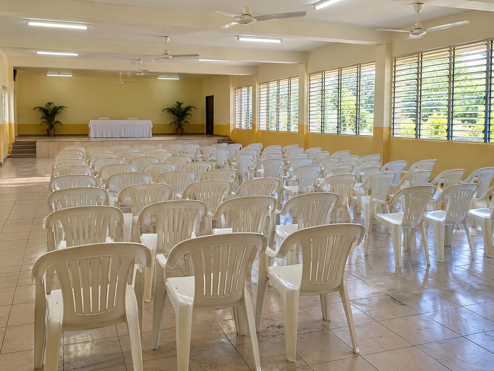 Rows of empty plain plastic chairs set in a community hall with louvred windows