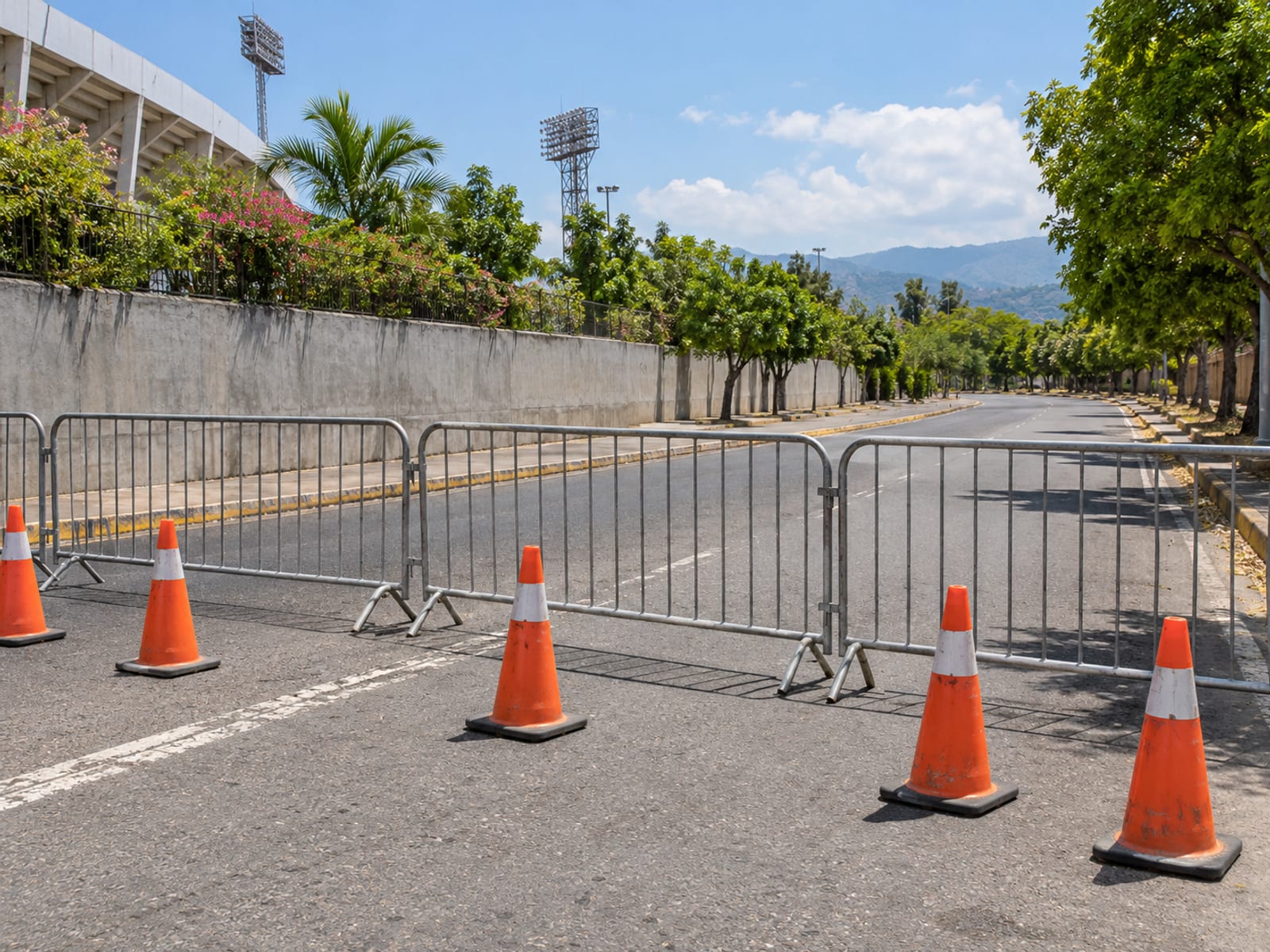Row of metal crowd barriers and orange traffic cones across a closed street in Kingston with trees and a wall behind
