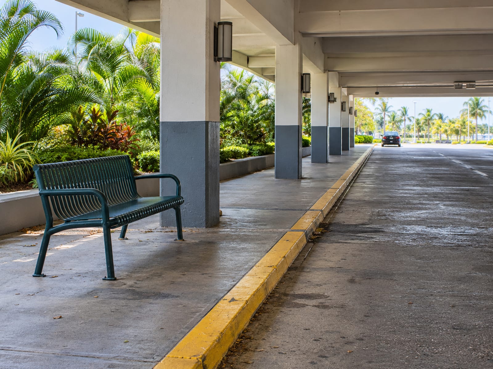 A covered concrete drop off bay with an empty bench and pillars, and one small plain car waiting at the far end.
