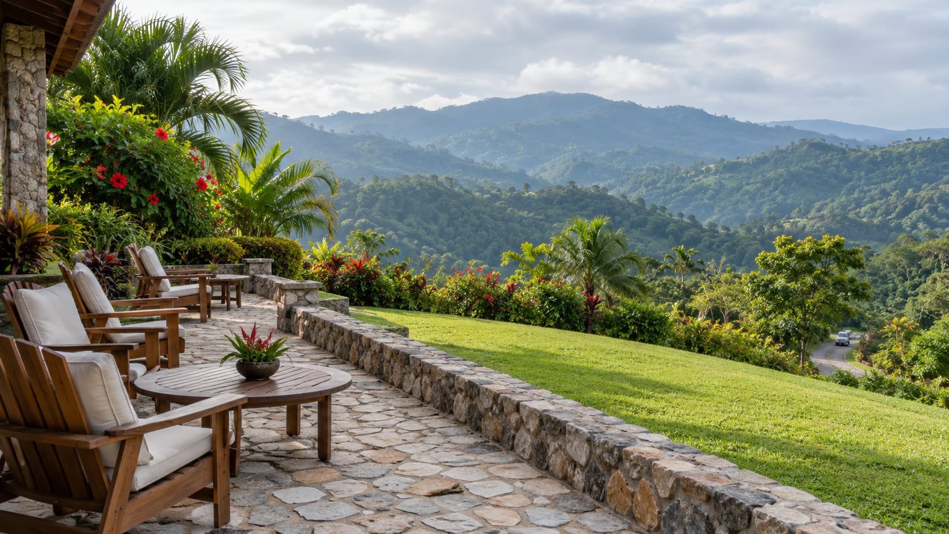 Wide view of an empty stone terrace with plain wooden chairs and a lawn running down to tree-covered hills under a pale morning sky