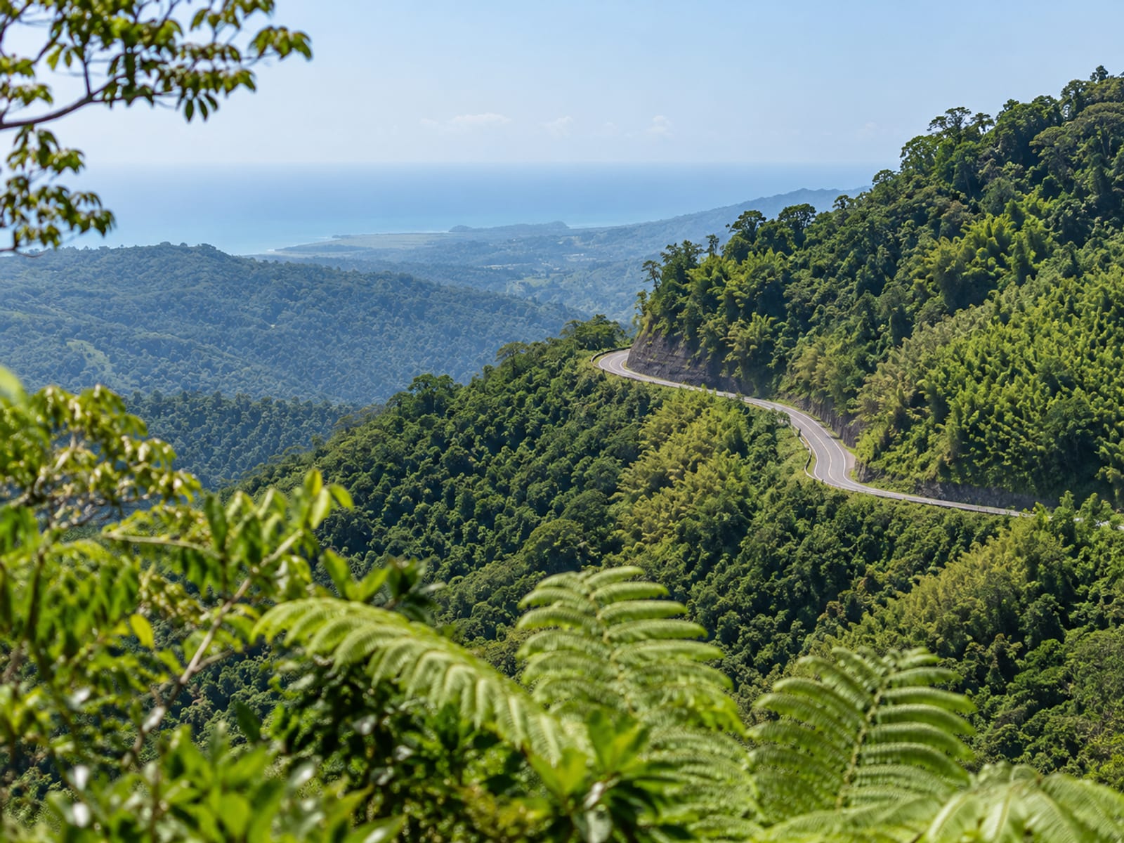 Narrow paved road winding along a green mountain ridge with a faint stretch of sea far in the distance