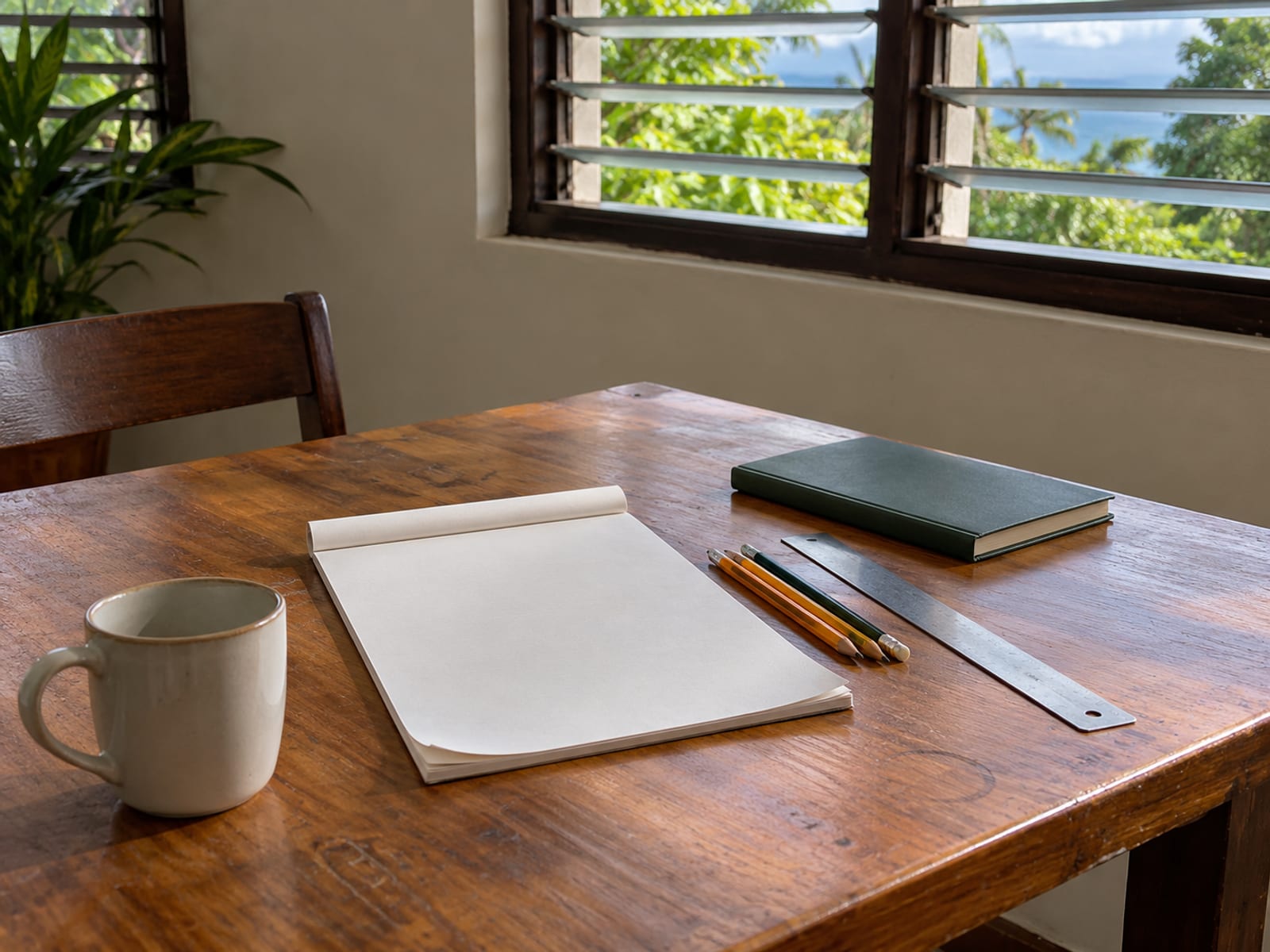 Wooden table by a window with a blank pad of paper, pencils, a ruler and an empty coffee mug