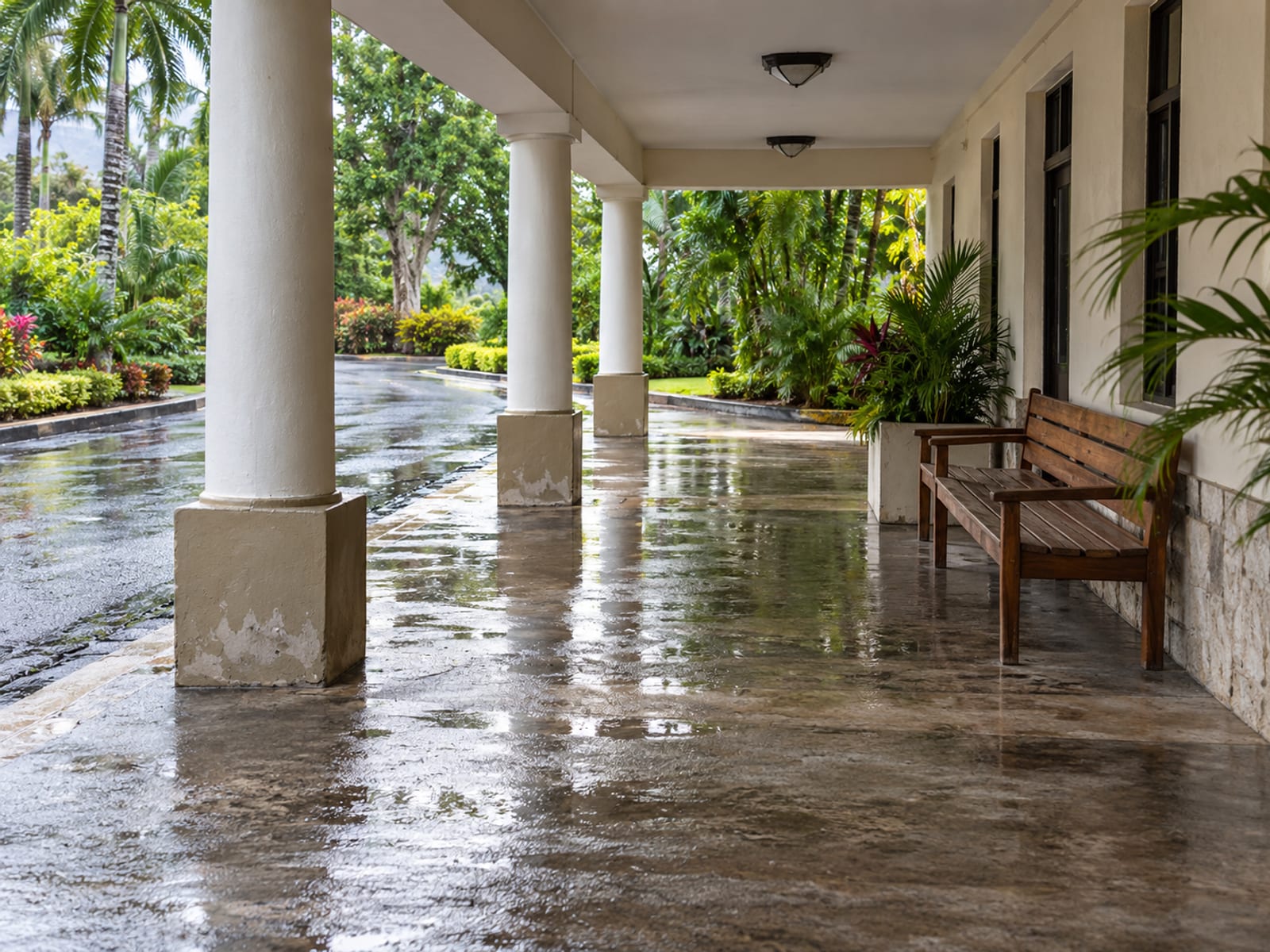Empty covered entrance bay of a plain painted building with concrete columns, a bench and a wet swept floor