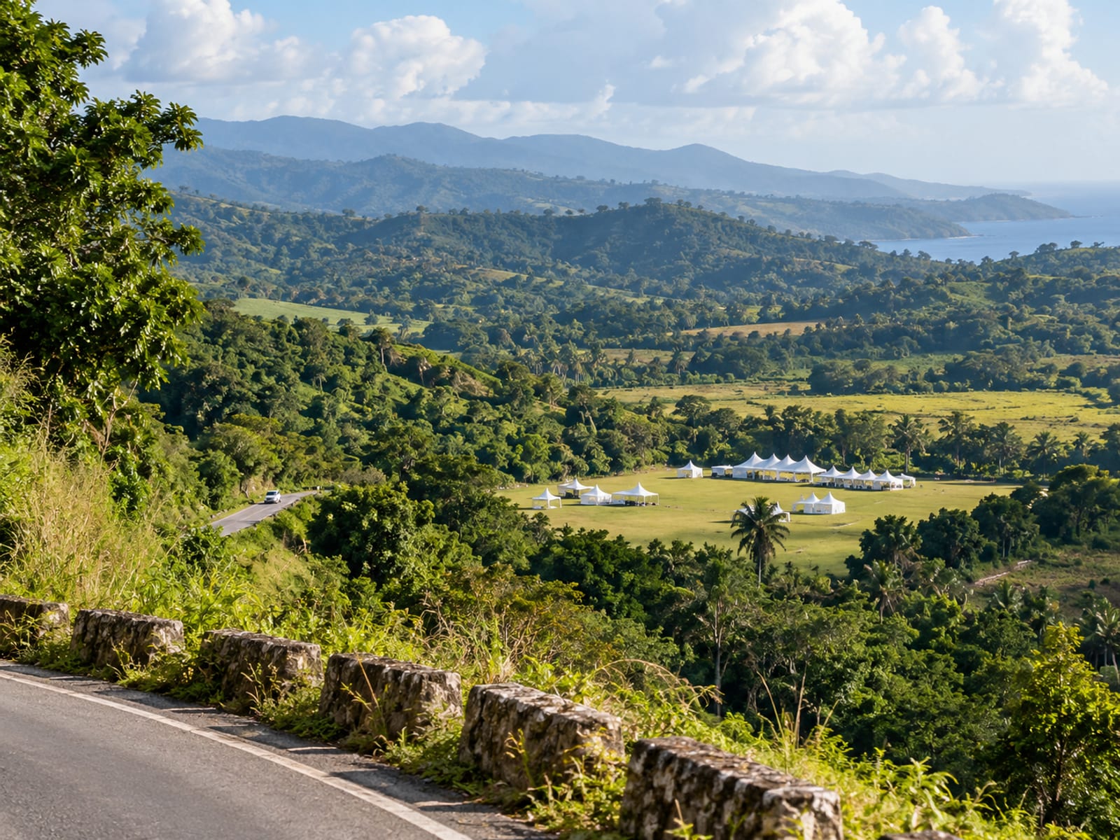 Distant valley with an open field and a few white tents in the hills seen from a ridge road