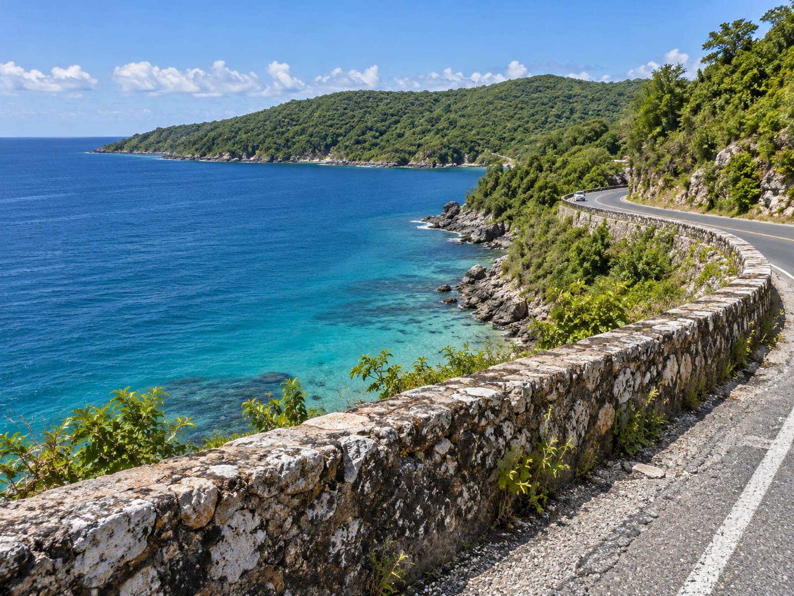 Coast road bend above a bay seen from a viewpoint with a small distant vehicle and a low wall