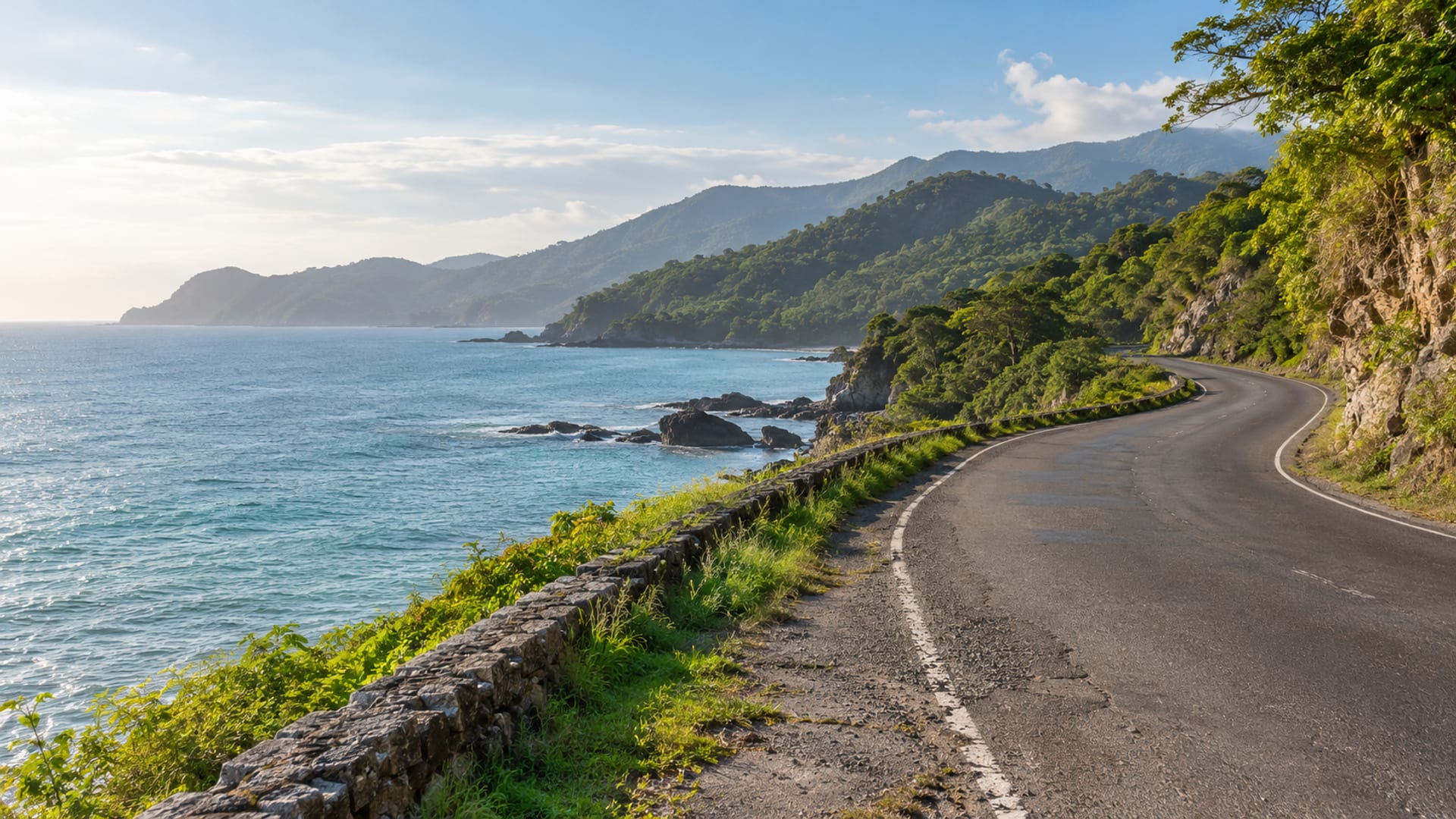 Empty two lane coastal road curving along a calm blue sea with green hills rising on the far side