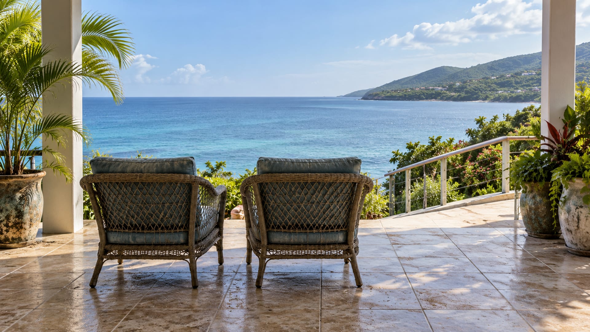 Two empty cushioned chairs on a level tiled verandah facing a calm sea, with a gentle ramp and potted plants
