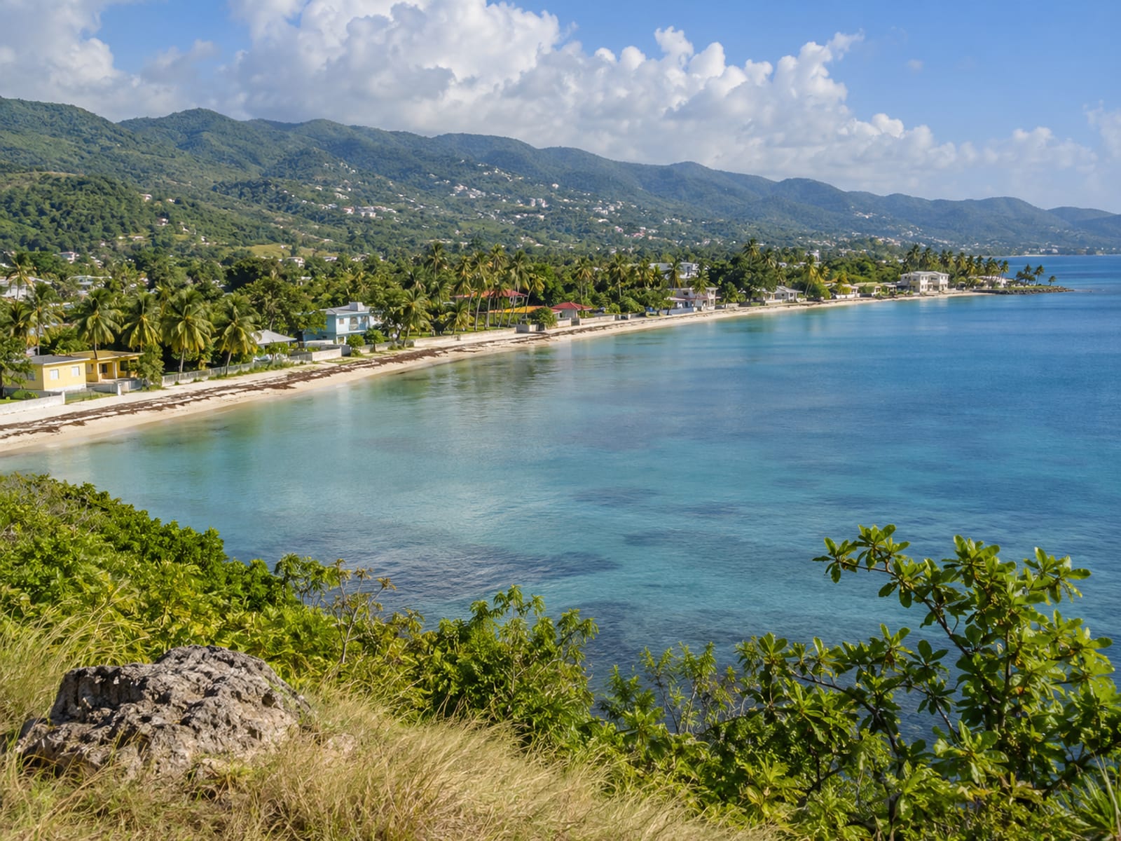 Wide view of a calm bay with low buildings along a flat shoreline and green hills behind