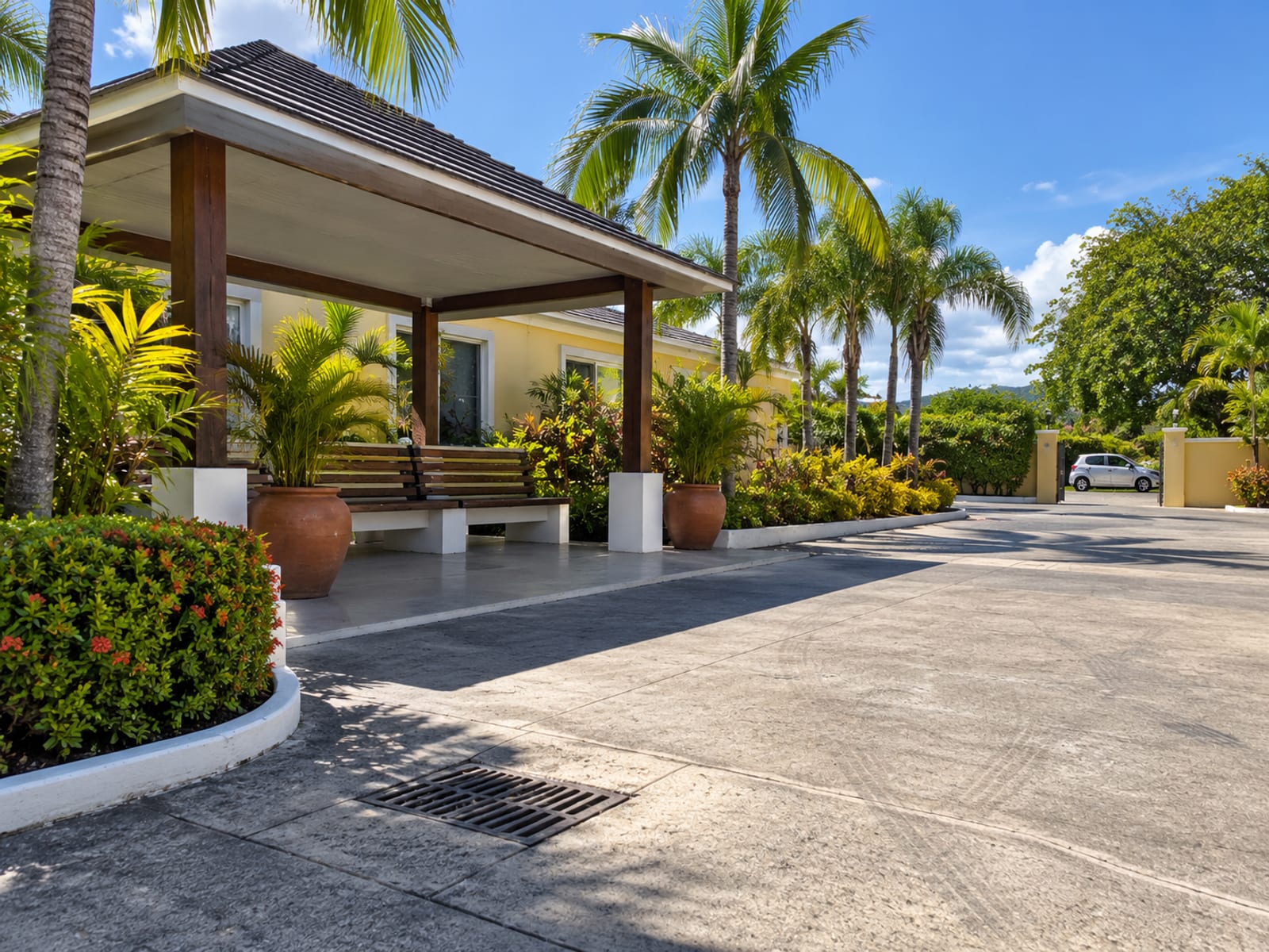 Empty paved forecourt with a roofed bench, palms and planters, one small car far near a gate