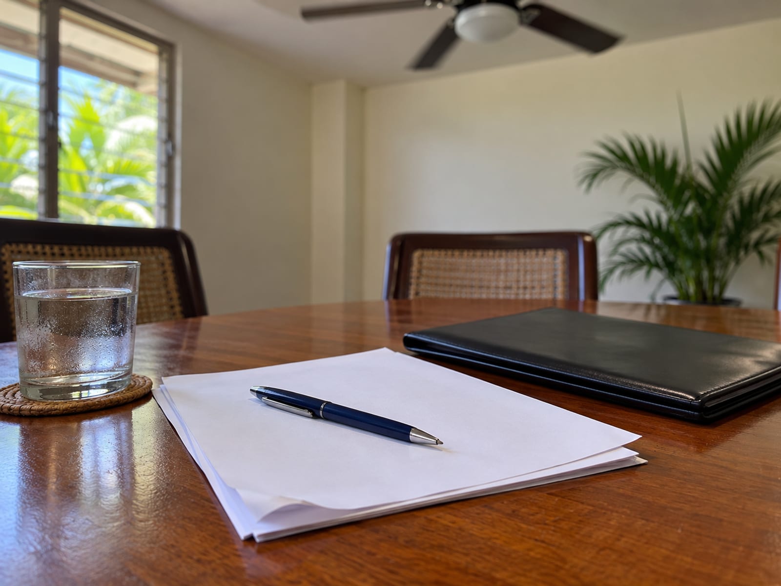Close view of blank pages, a pen and a closed folder on a table with a ceiling fan blurred above