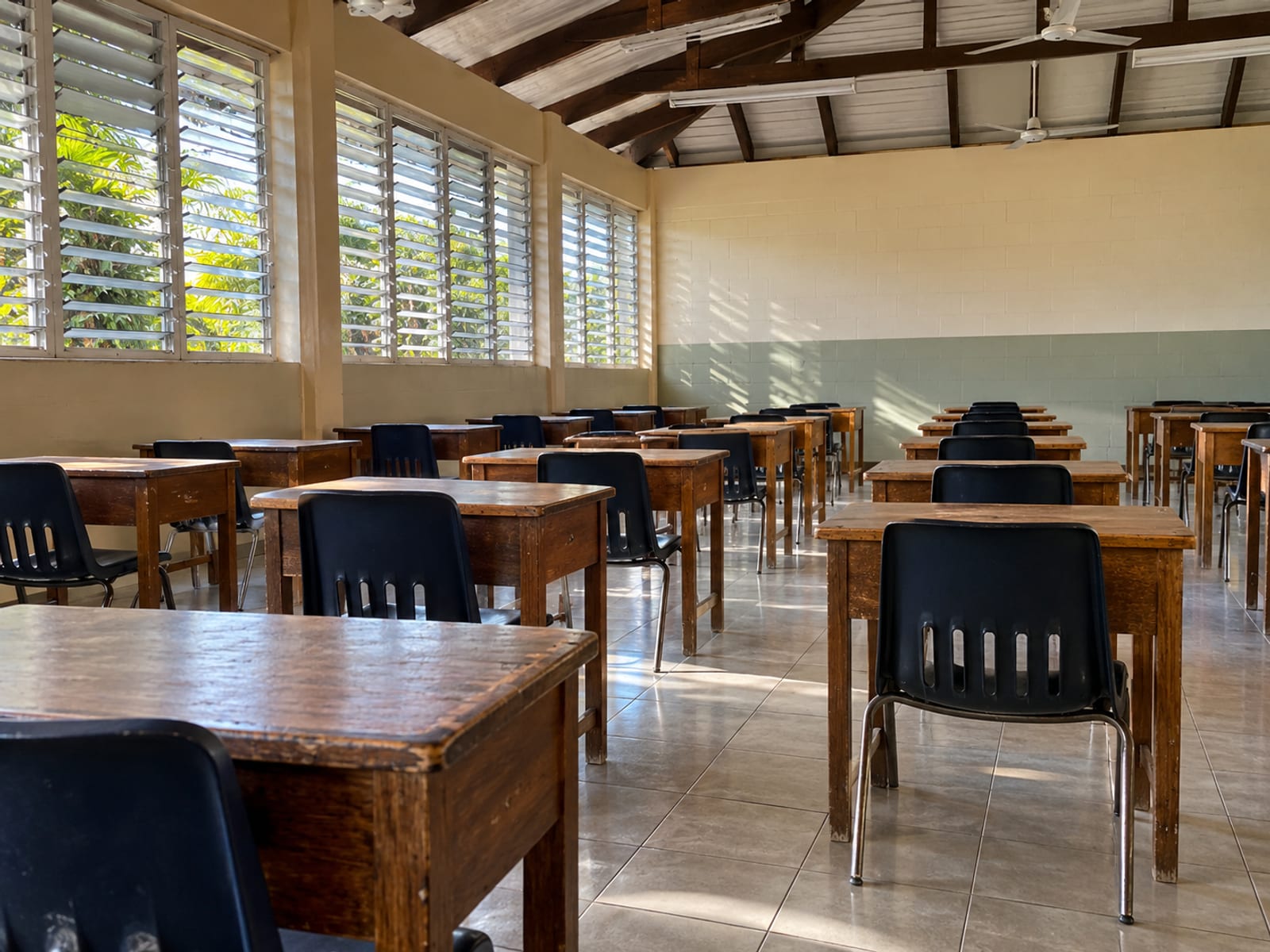 Rows of empty wooden desks and chairs spaced apart in a plain hall with louvre windows and a bare floor