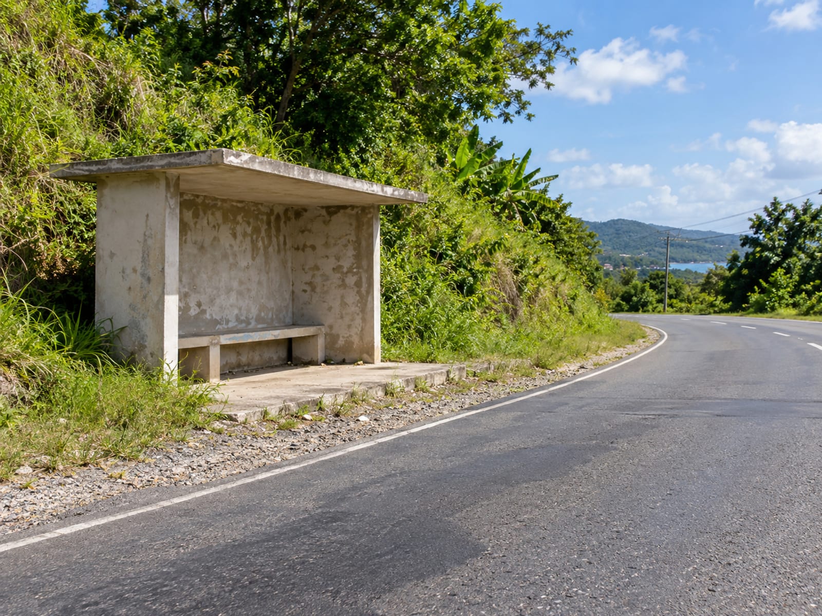 Plain open concrete bus shelter with a bench beside a rural road, empty, with green bank behind