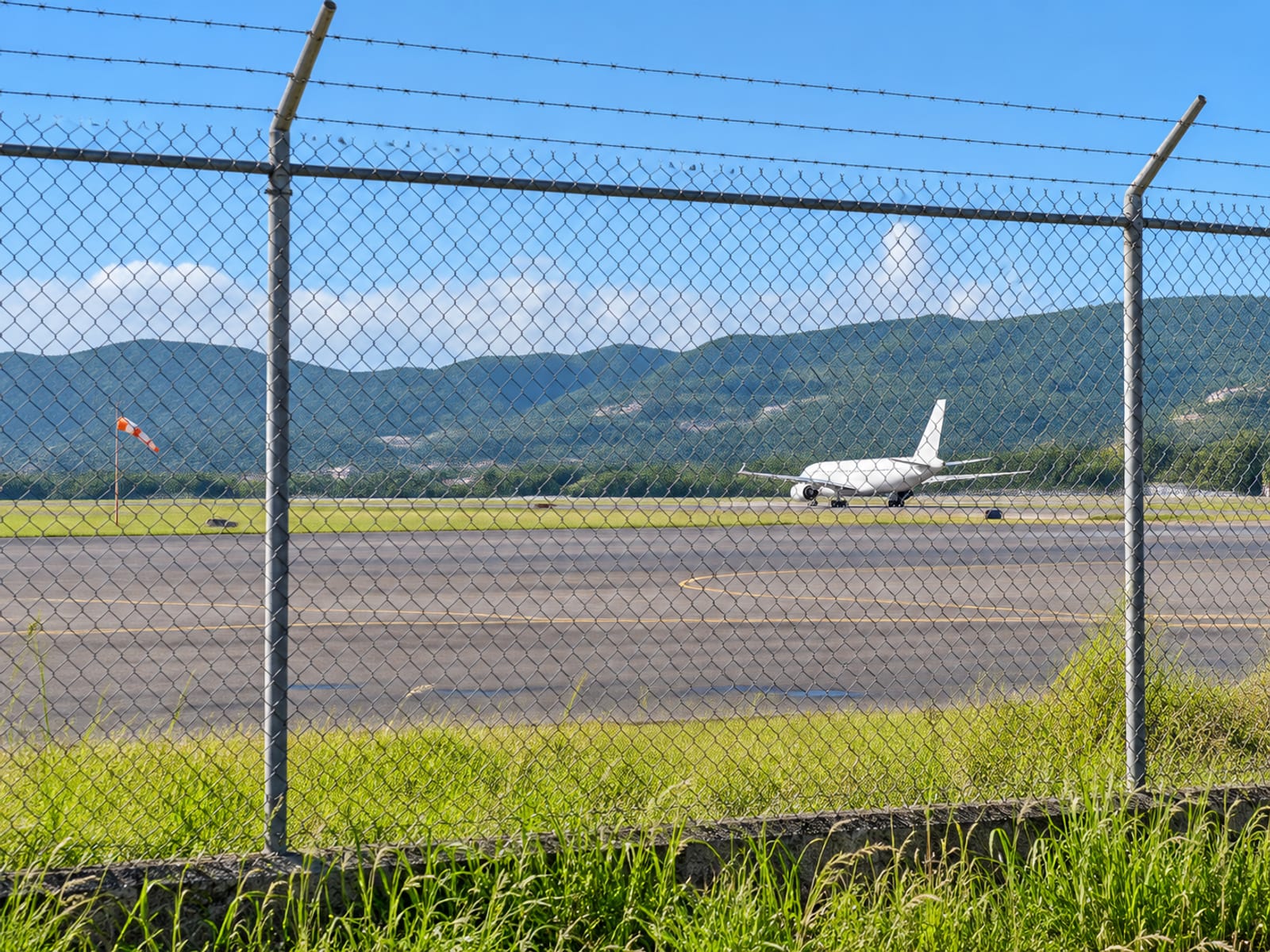 Distant view across an airport tarmac toward a plain white aircraft with green hills behind