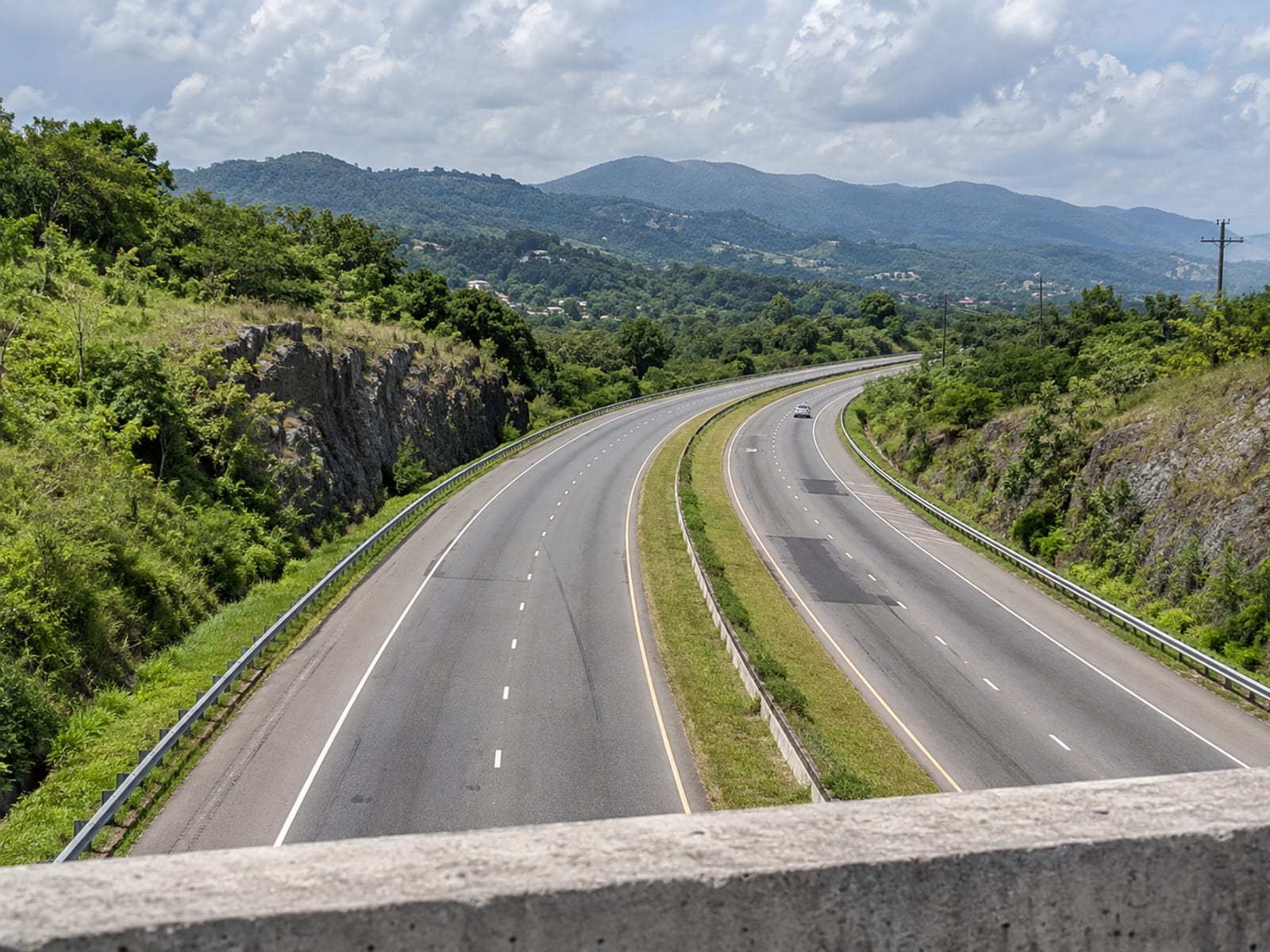Divided highway curving through green hills seen from an overpass, with one small vehicle far away