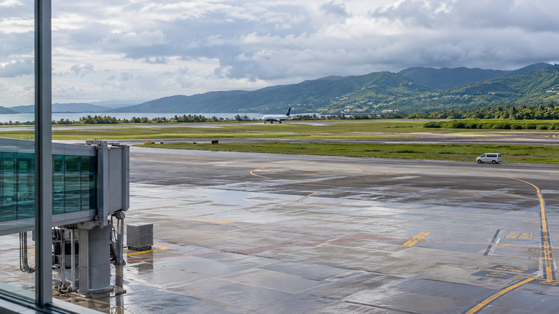 Wide view across an airport apron to a runway and green hills beyond, with one small distant aircraft under soft grey cloud