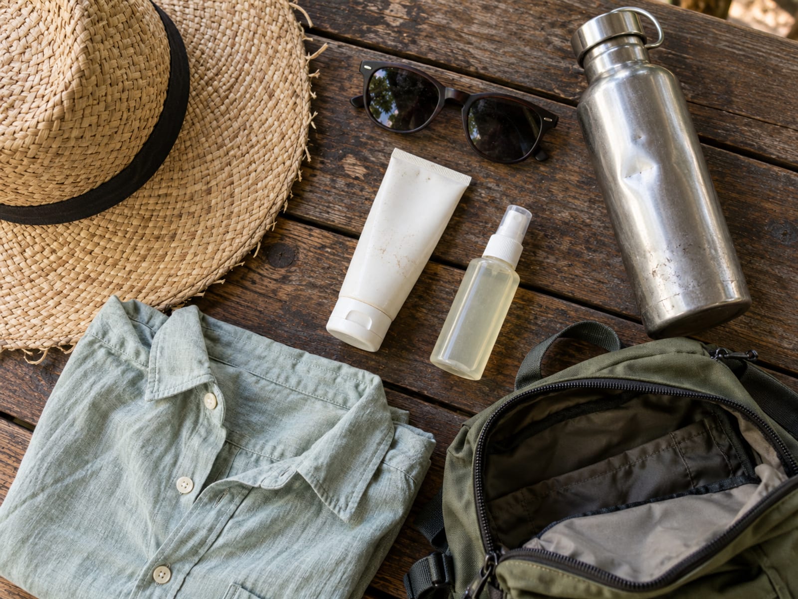 A straw sun hat, sunglasses, a plain steel bottle and an unlabelled sun cream tube laid on a wooden table beside an open small backpack
