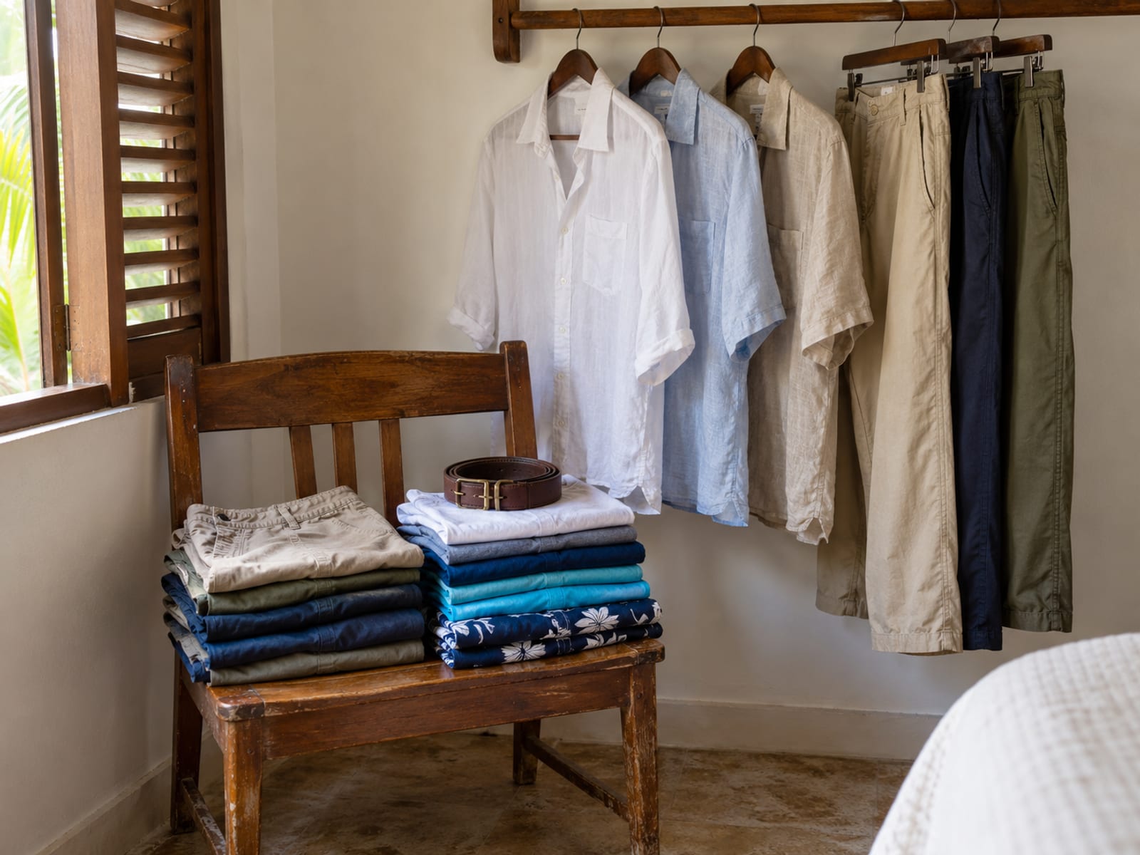 Rail with light shirts and long trousers on hangers, with folded shorts and T-shirts stacked on a chair below