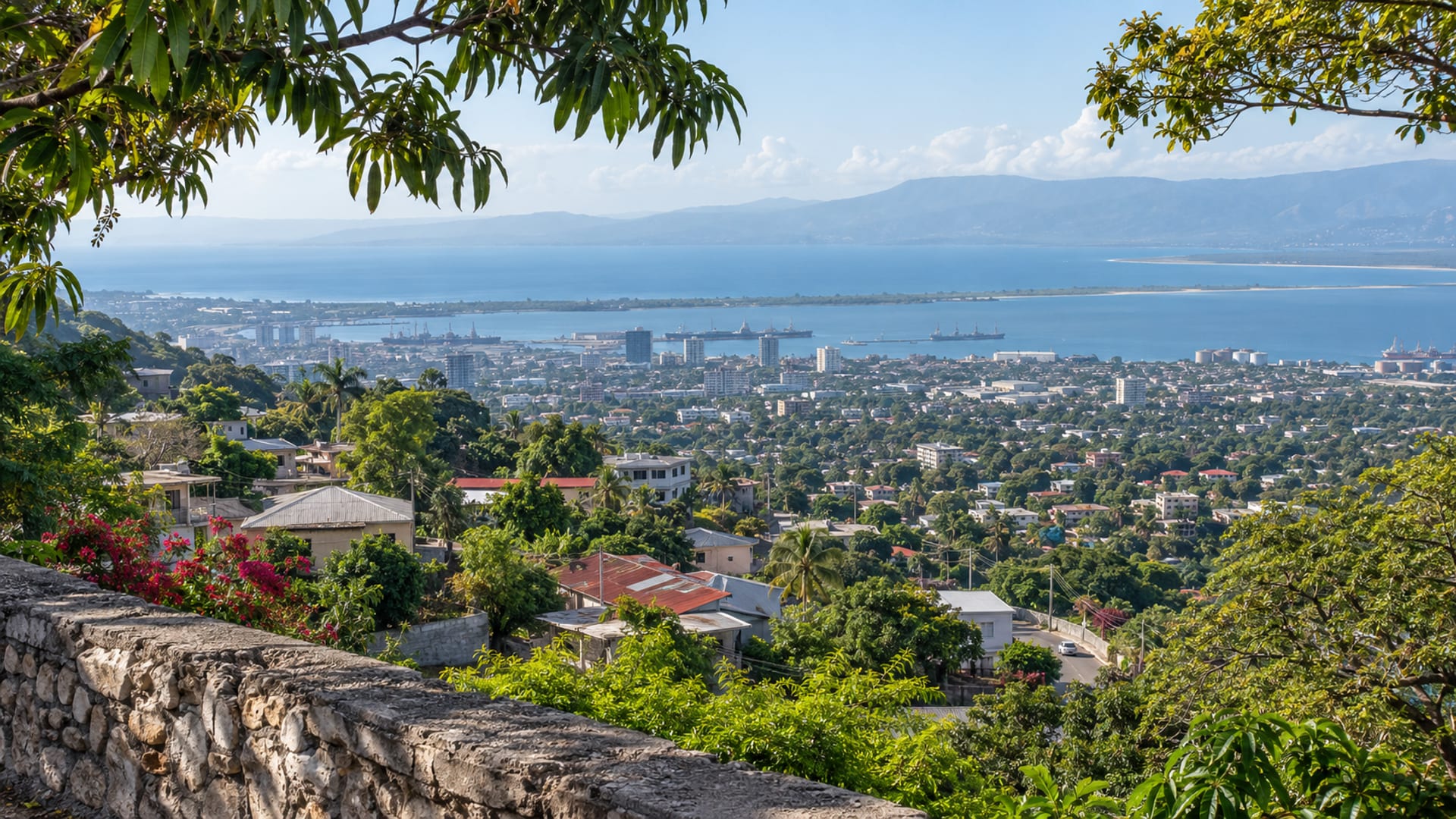 Wide view from a green hillside across Kingston rooftops toward a hazy harbour and long sandspit under a pale blue sky, with one tiny vehicle on a distant road.