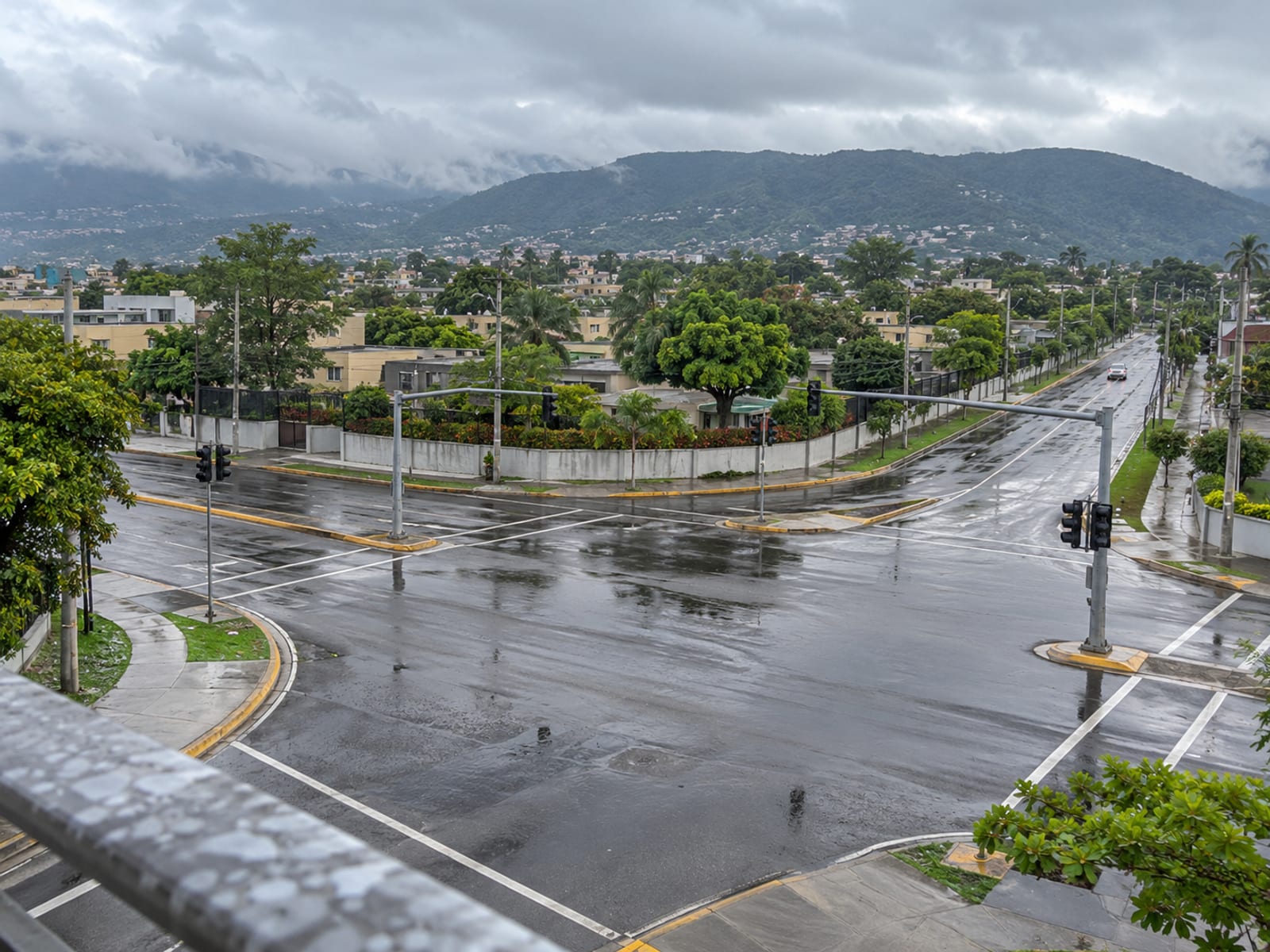 Empty wet road junction seen from above under a heavy grey sky, with traffic lights, shining tarmac and one small distant vehicle.