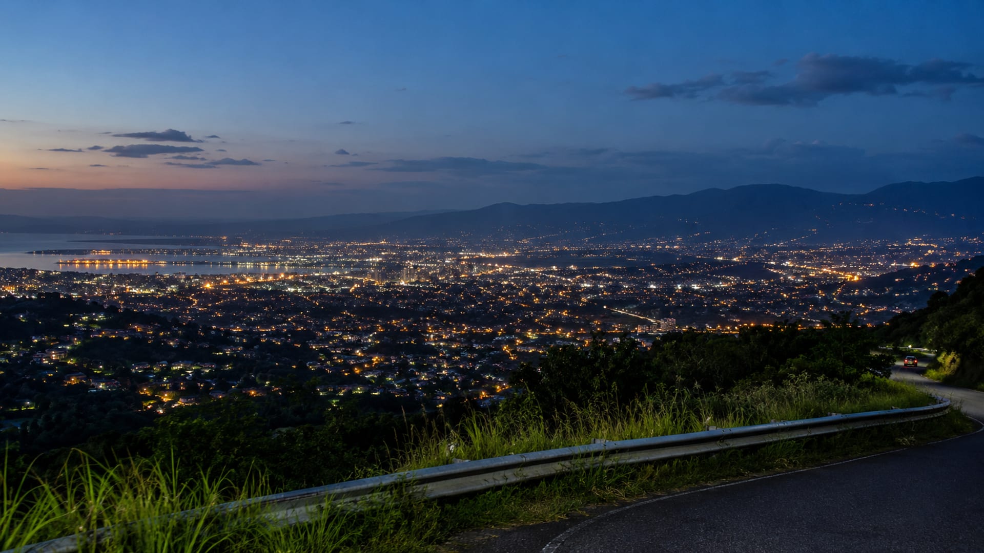 Wide dusk view from a ridge across scattered Kingston lights under a deep blue sky, with a faint ribbon of road below.