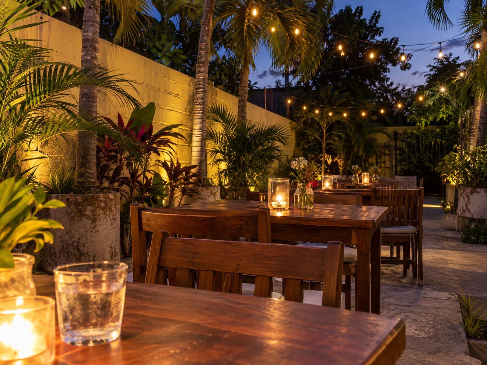 Courtyard with empty wooden tables, lit candles in glass jars, string lights overhead and potted palms in blue dusk.