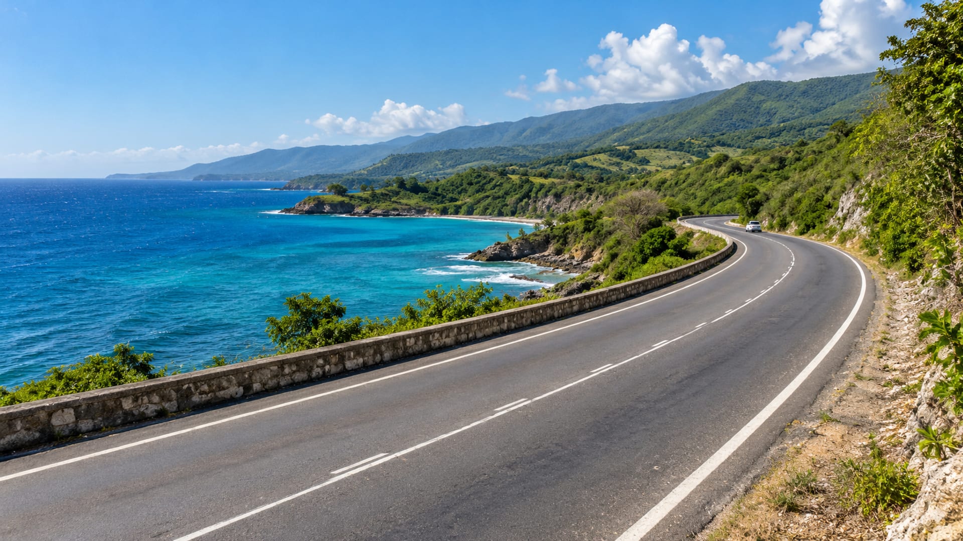 Wide paved coastal highway curving beside blue sea with one small distant car and green hills inland.