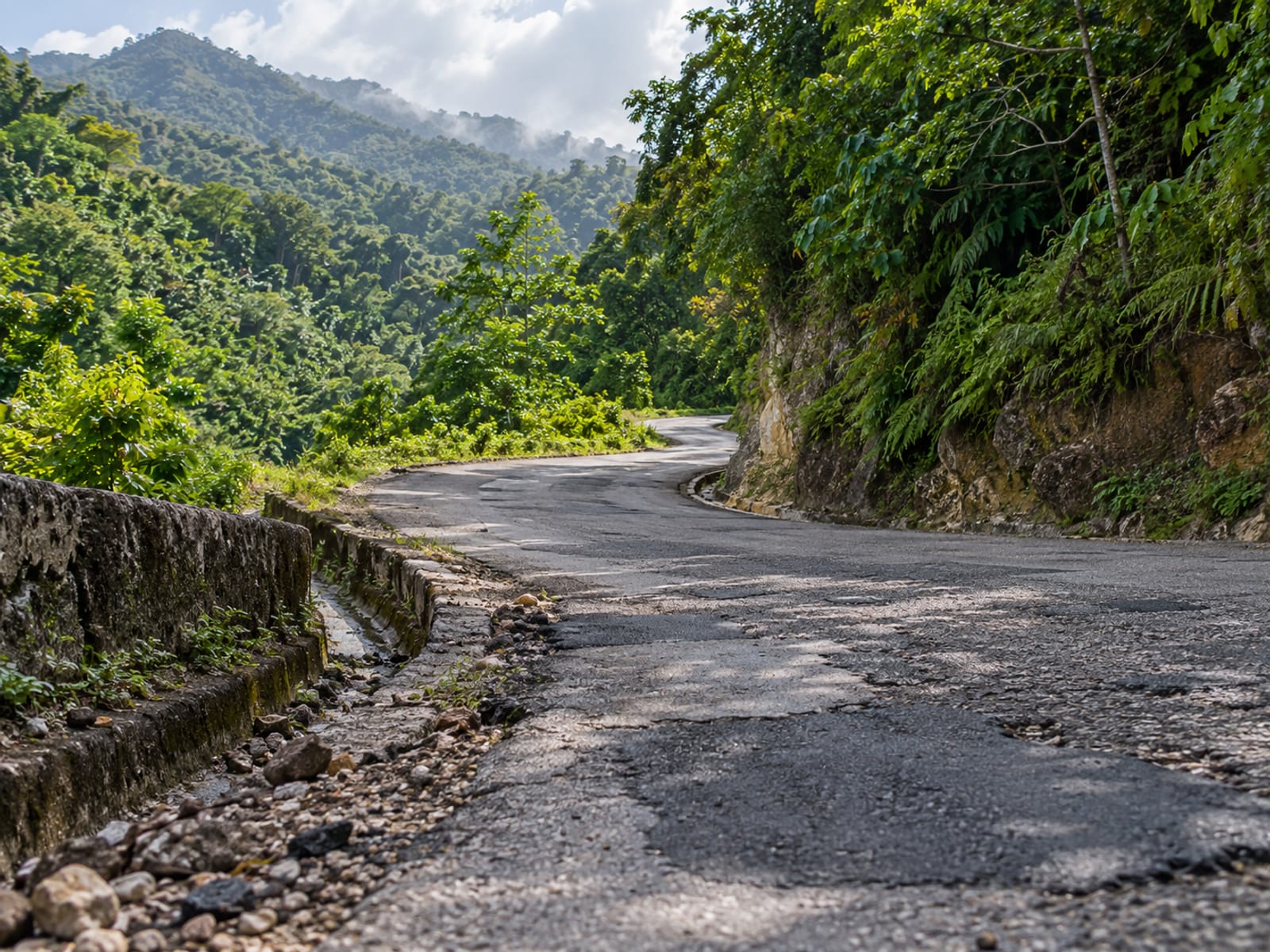 Narrow patched asphalt road winding through steep green hills with a rough verge.