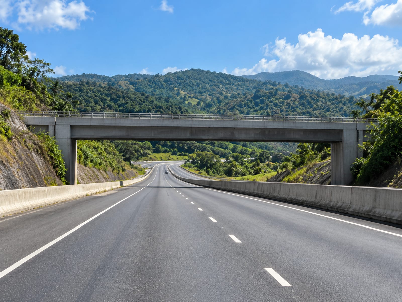 Wide empty multi-lane motorway passing under a concrete overpass with green hills beyond.