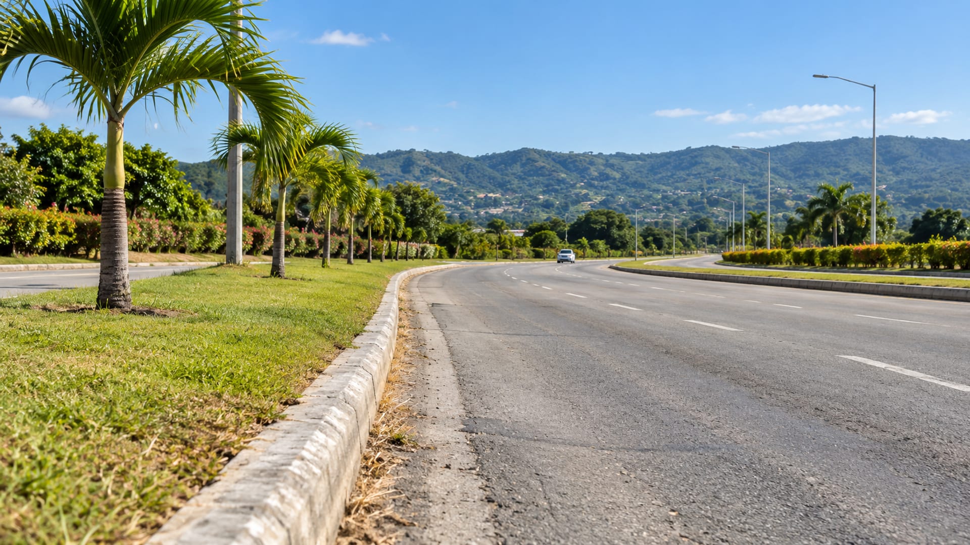 Wide view of a quiet airport approach road with a plain kerb, young palms and green hills behind under a bright morning sky