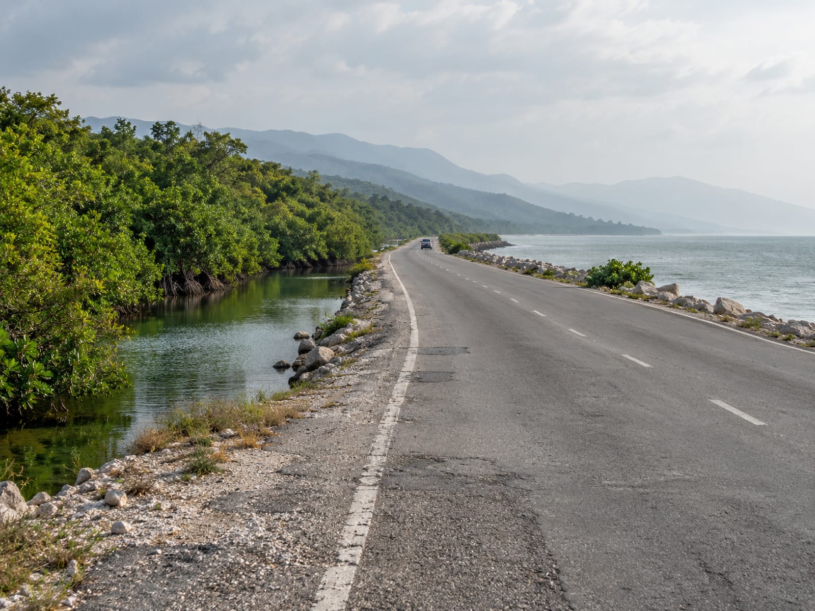 Two lane coastal road running between mangrove trees and calm grey green sea under hazy sky