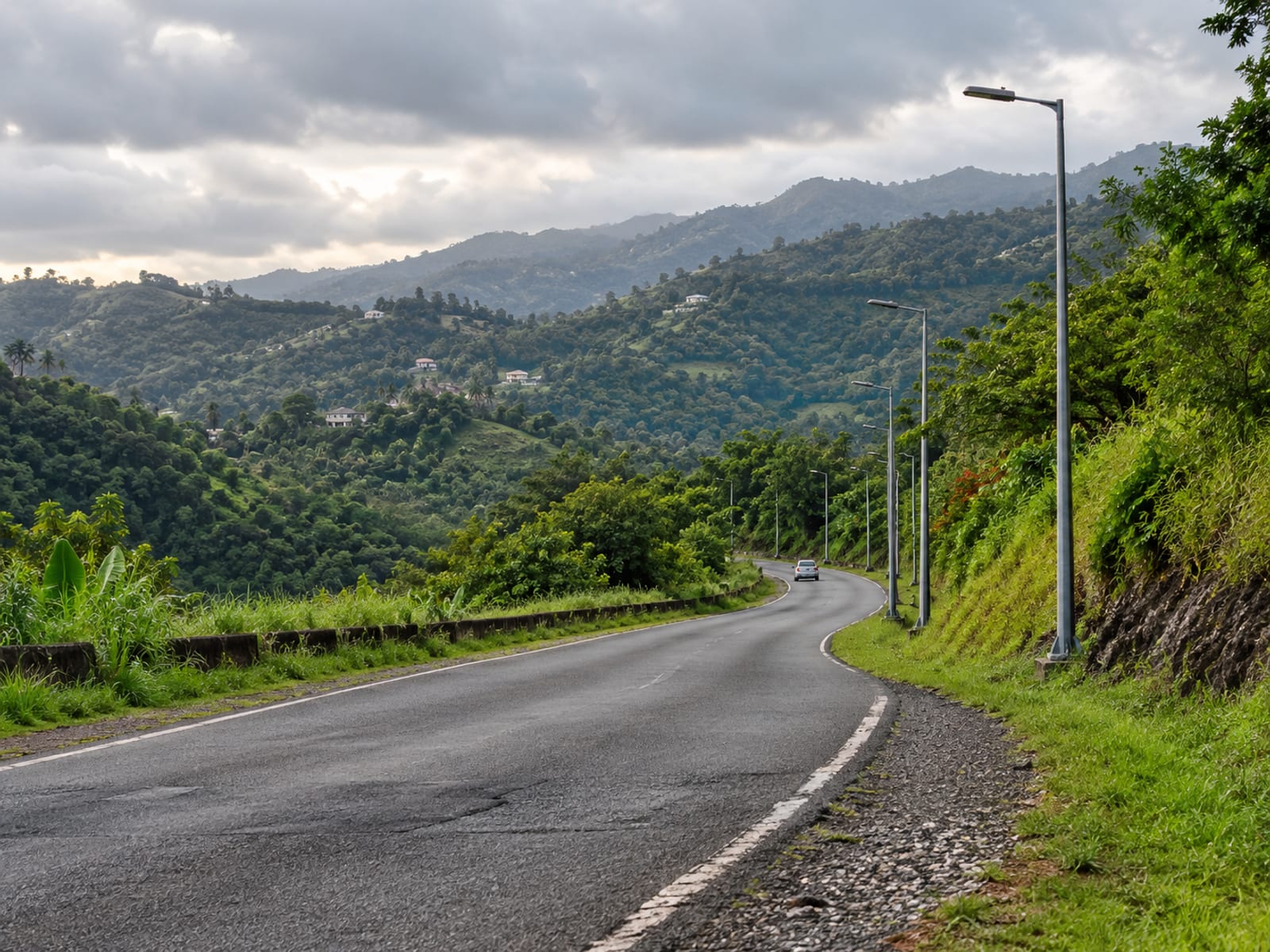 Wide view of a quiet country road bending past a row of unlit lamp posts and a grass bank, one tiny plain car far away