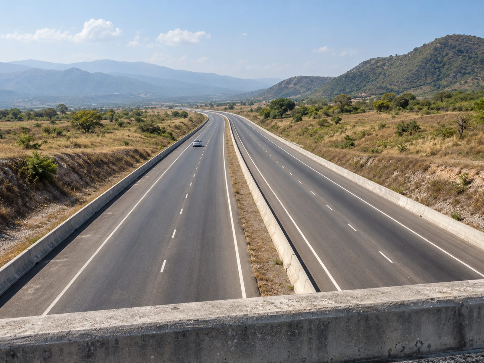 Modern divided highway crossing flat dry land near hills seen from a bridge, with one distant car