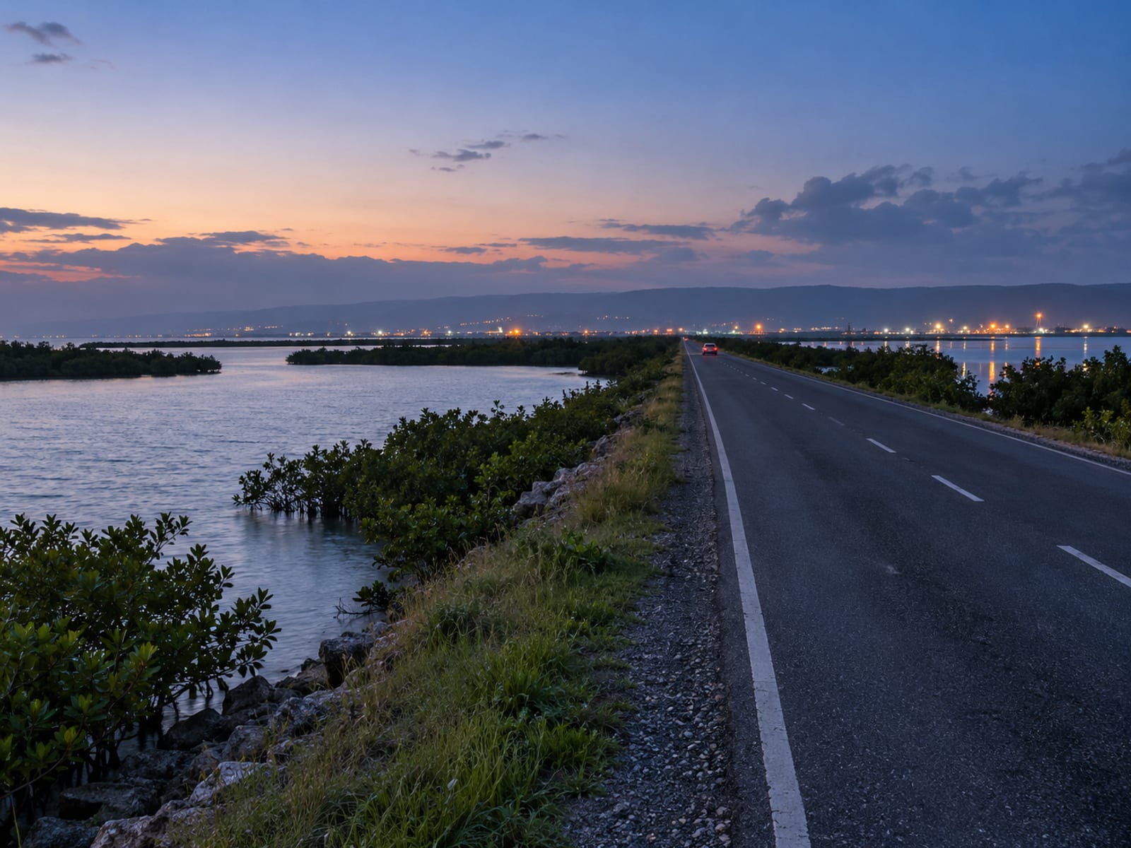 Long straight road across flat land beside a water inlet before dawn with distant orange lights