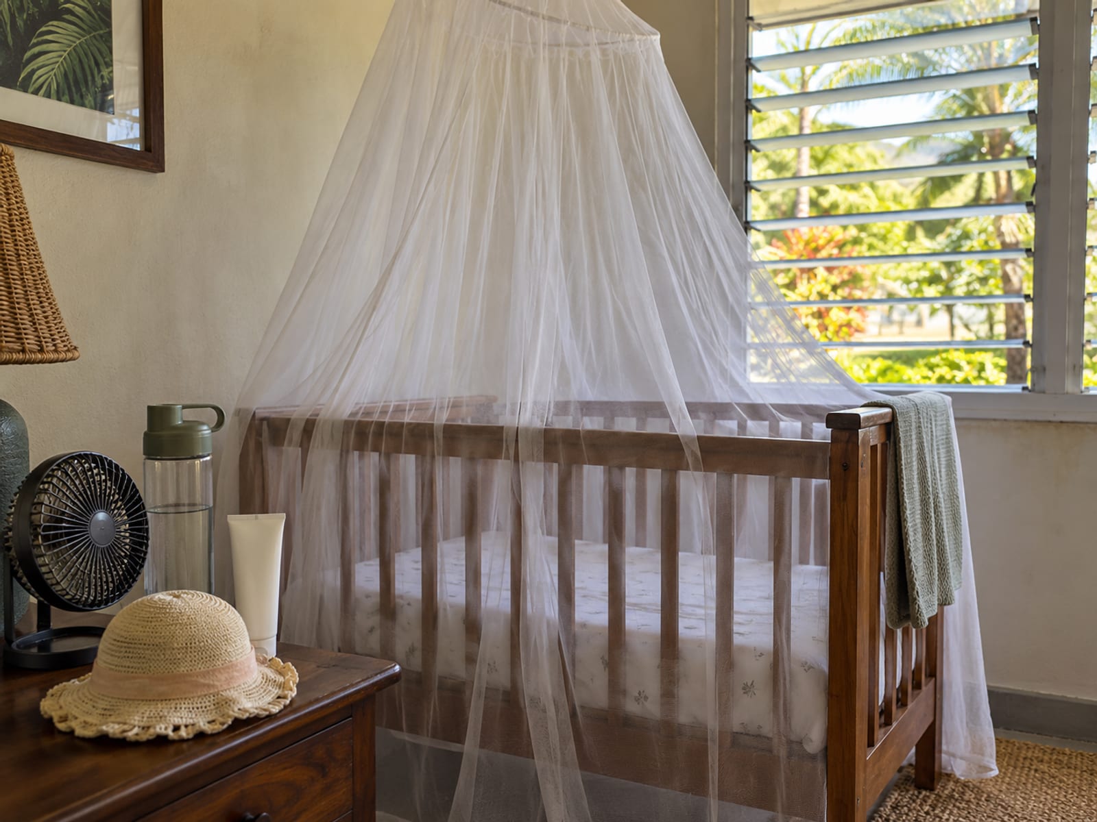 Cot draped with a fine mosquito net beside a small sun hat and a blank sunscreen tube on a side table in a shaded room