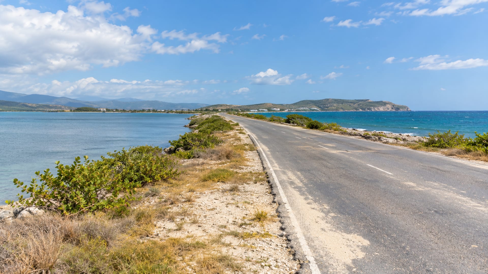 Long straight coastal road on a narrow sandy peninsula with sea on both sides and low hills far ahead