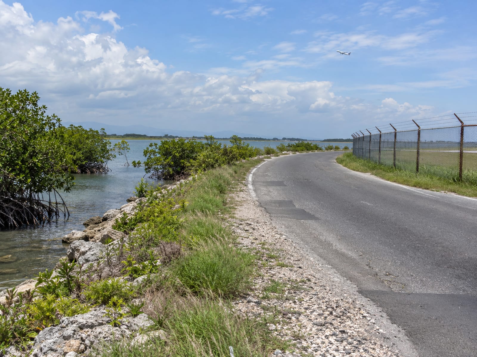 Roadside view of a long perimeter fence, mangrove edge and a tiny distant aircraft in the sky beyond the water