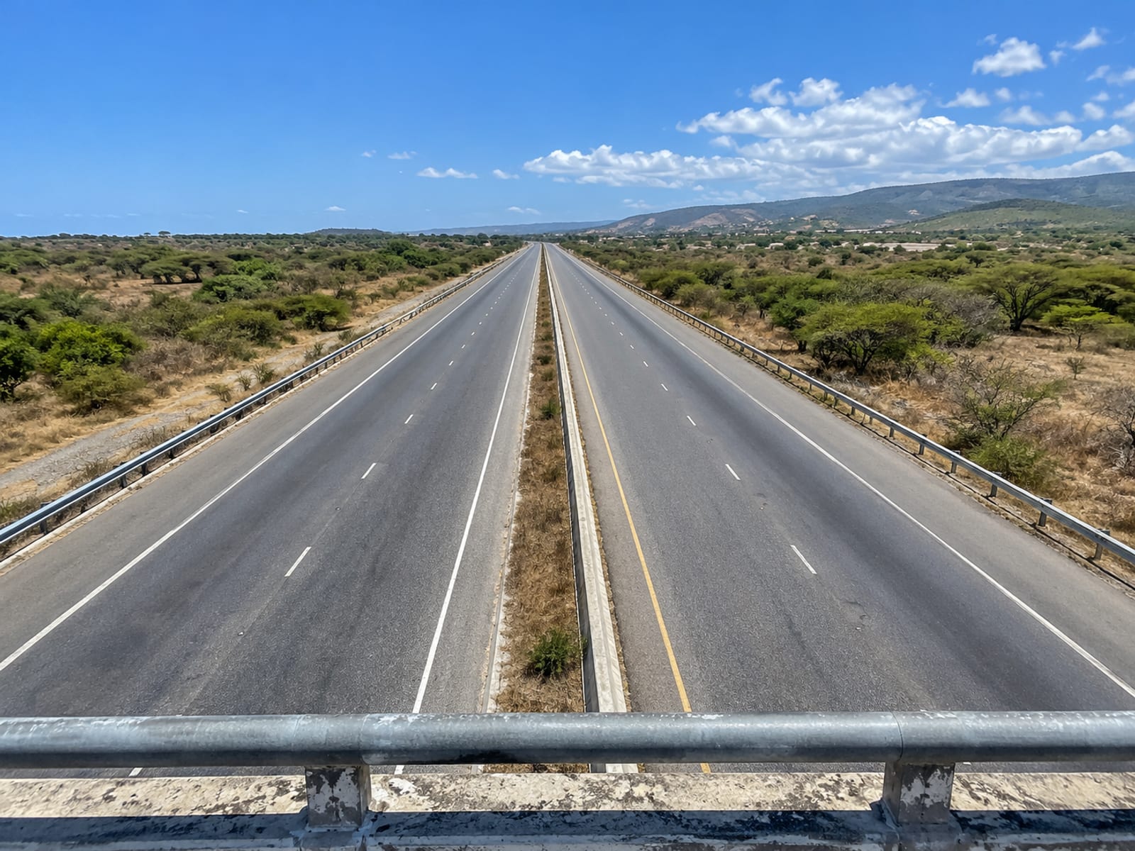 Elevated view along an empty divided highway running straight through dry scrub and low trees under a bright sky.