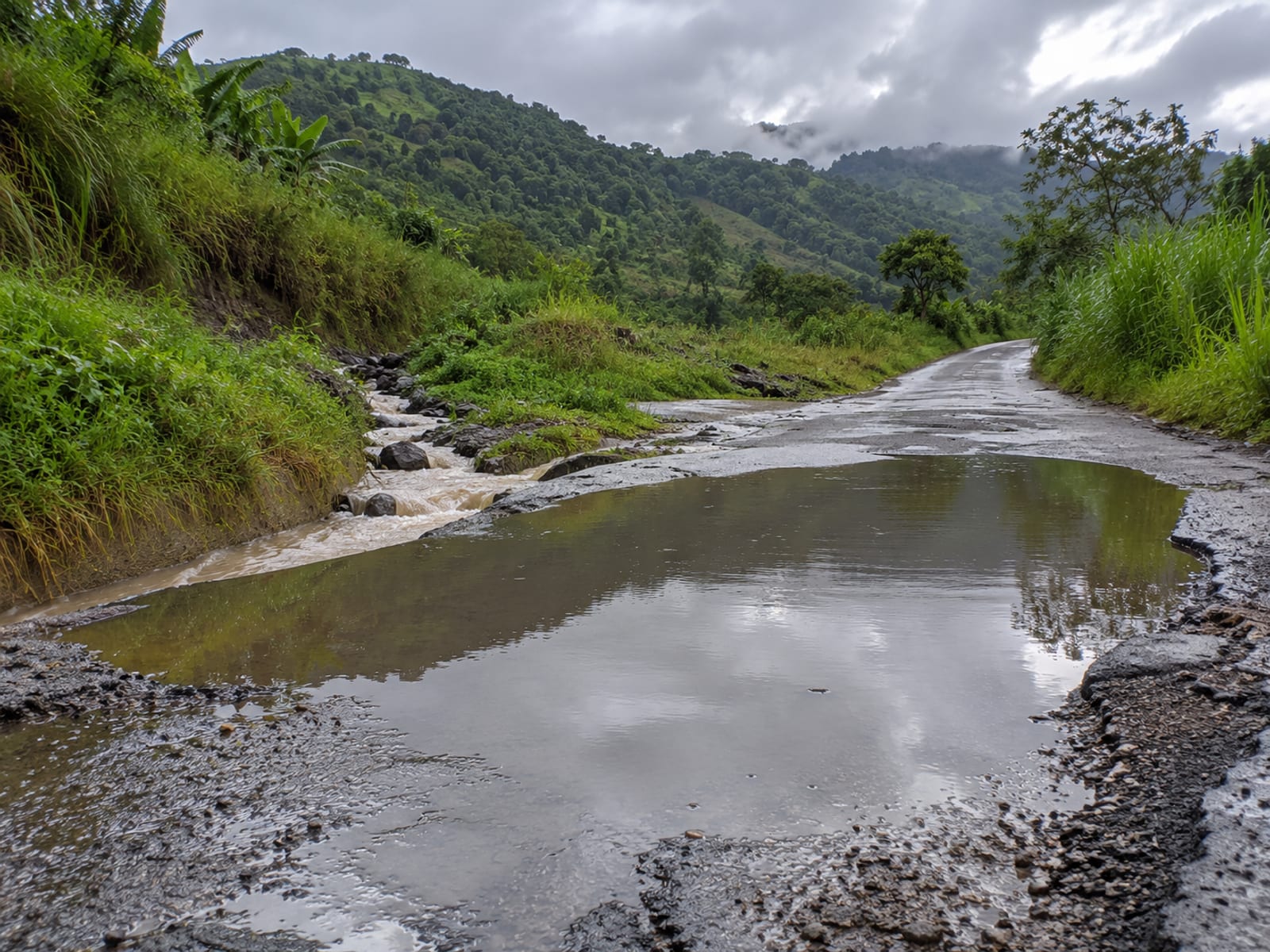 Rural road after rain with puddles in a dip, a roadside gully and green hills under heavy cloud
