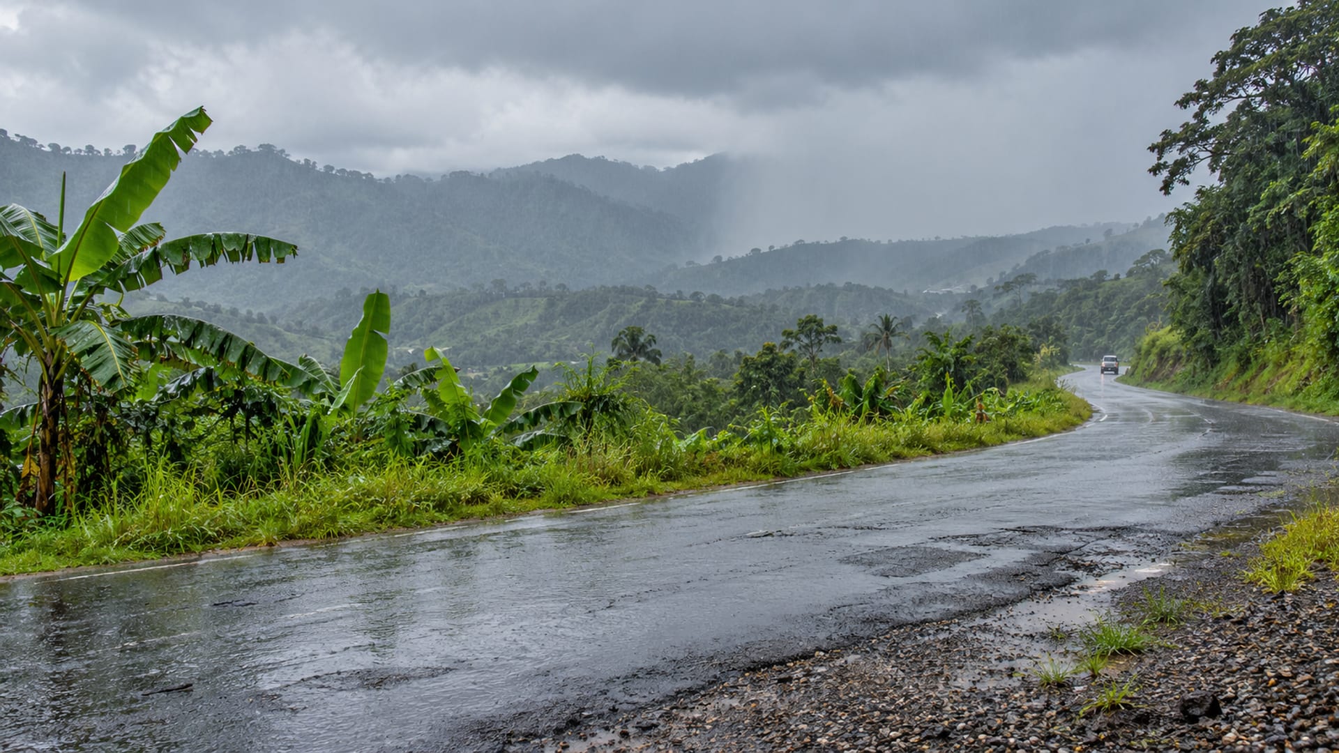Wet road beside green hills under heavy grey rain clouds with rain falling over the distance