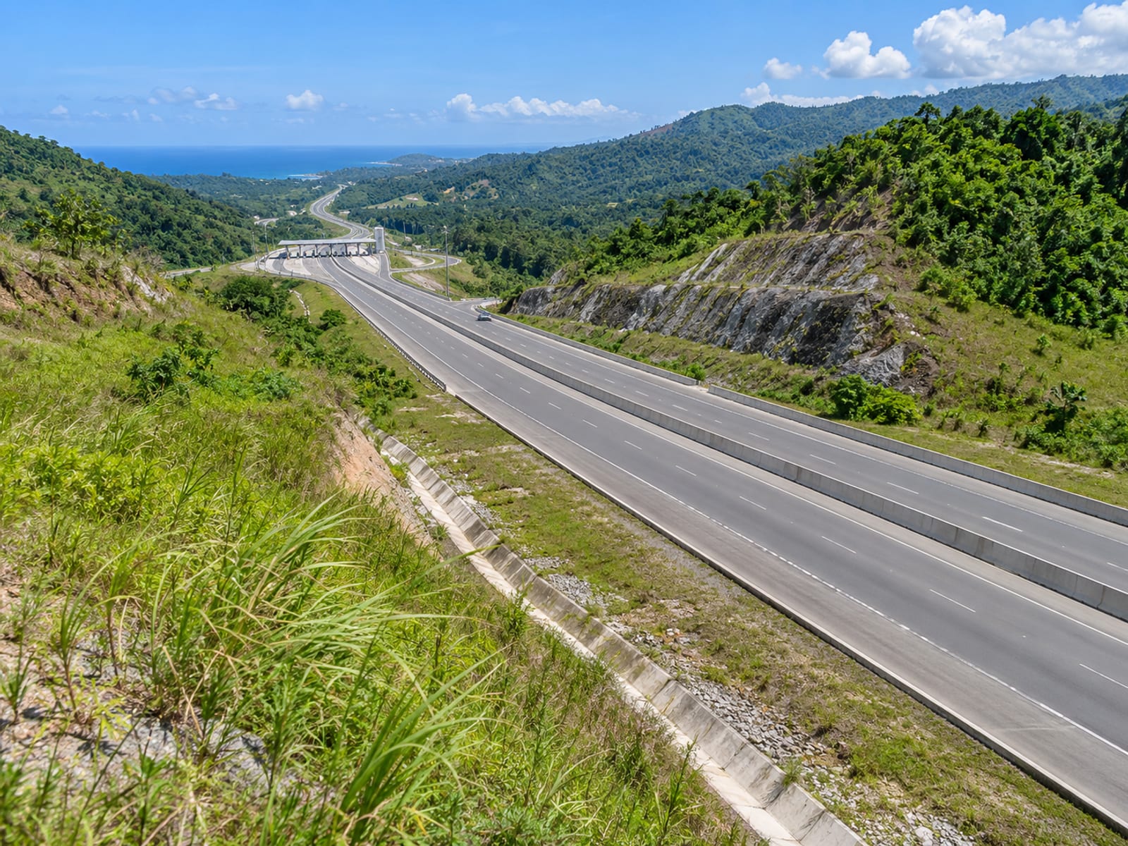 Multi lane highway cutting through green hills seen from a high bank with a tiny plaza in the distance