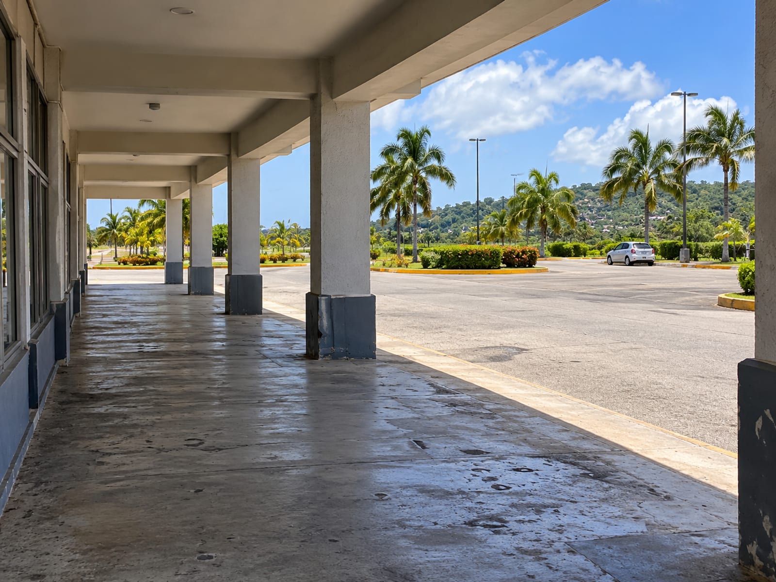 A covered concrete walkway with plain pillars running beside an empty paved car park under bright sky.