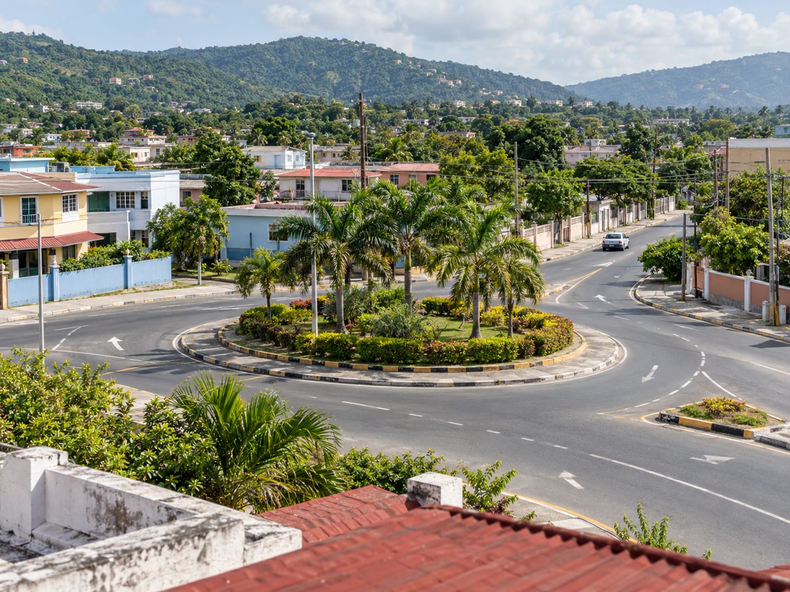 Elevated view of a small town roundabout with palm trees and low painted buildings, with one tiny plain car on the road.