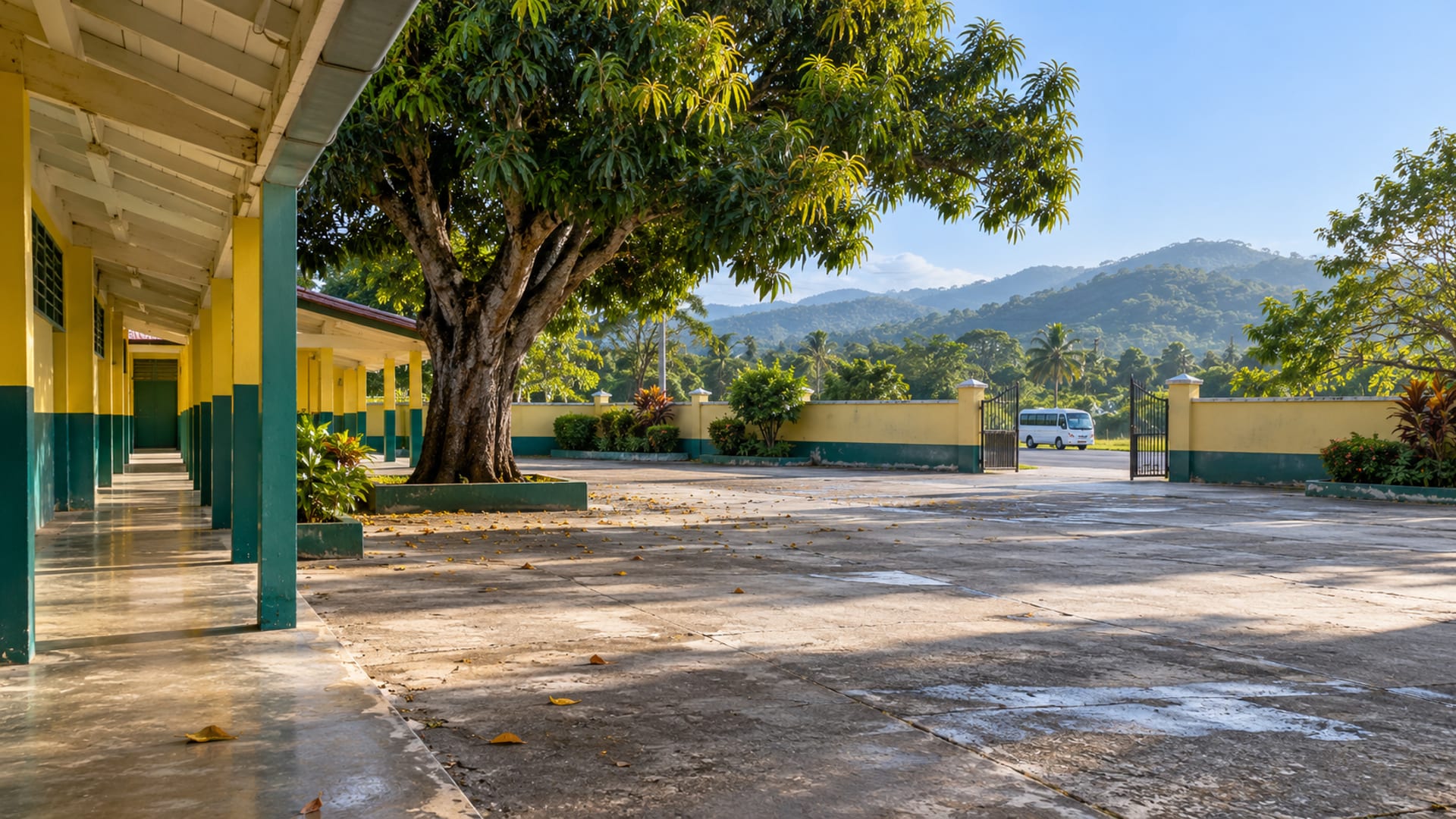 Empty concrete schoolyard with a covered walkway and a mango tree, green hills beyond the fence and one small plain minibus far in the distance