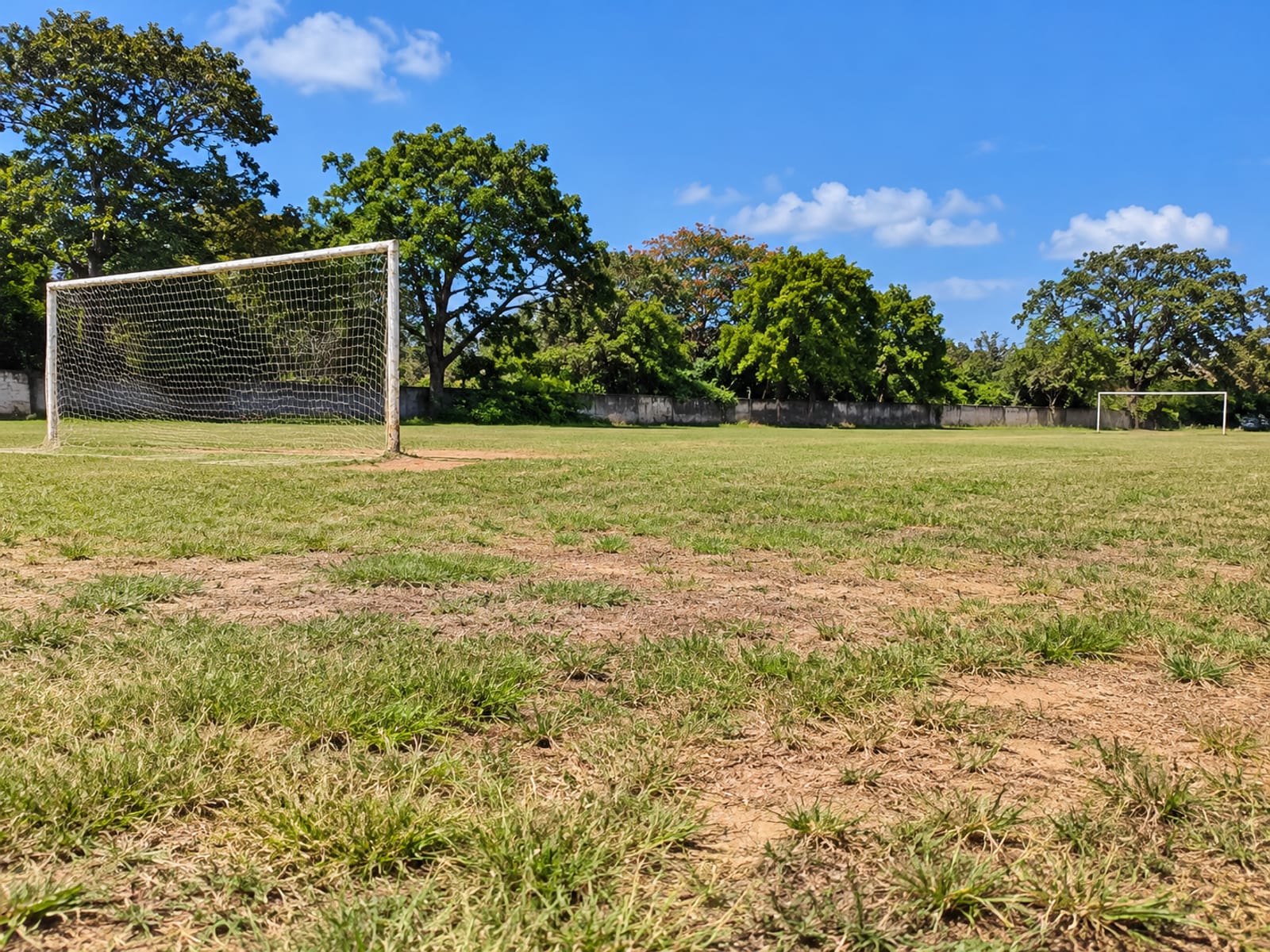 An empty grassy school playing field with two simple goalposts, trees along the boundary and a low wall in the distance.