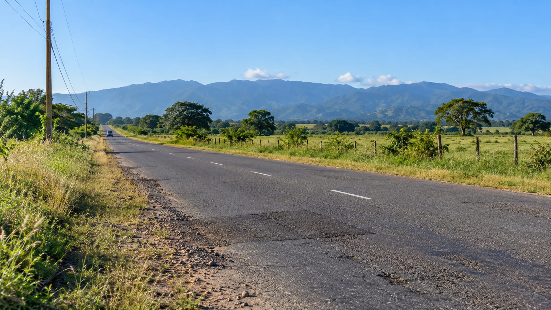 Wide view of a straight road crossing a flat green plain with scattered trees and blue hills in the far distance.