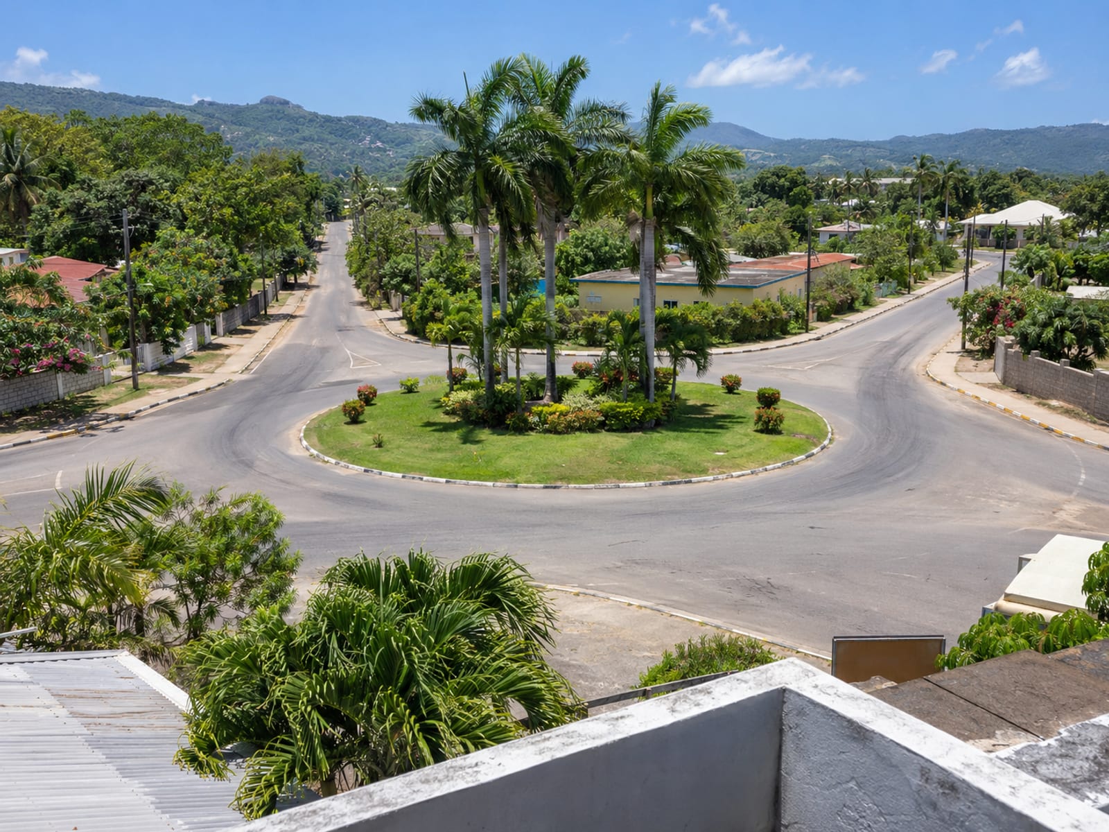 Raised view of an empty roundabout with a planted centre of palms where three roads meet, with low buildings at the edge.